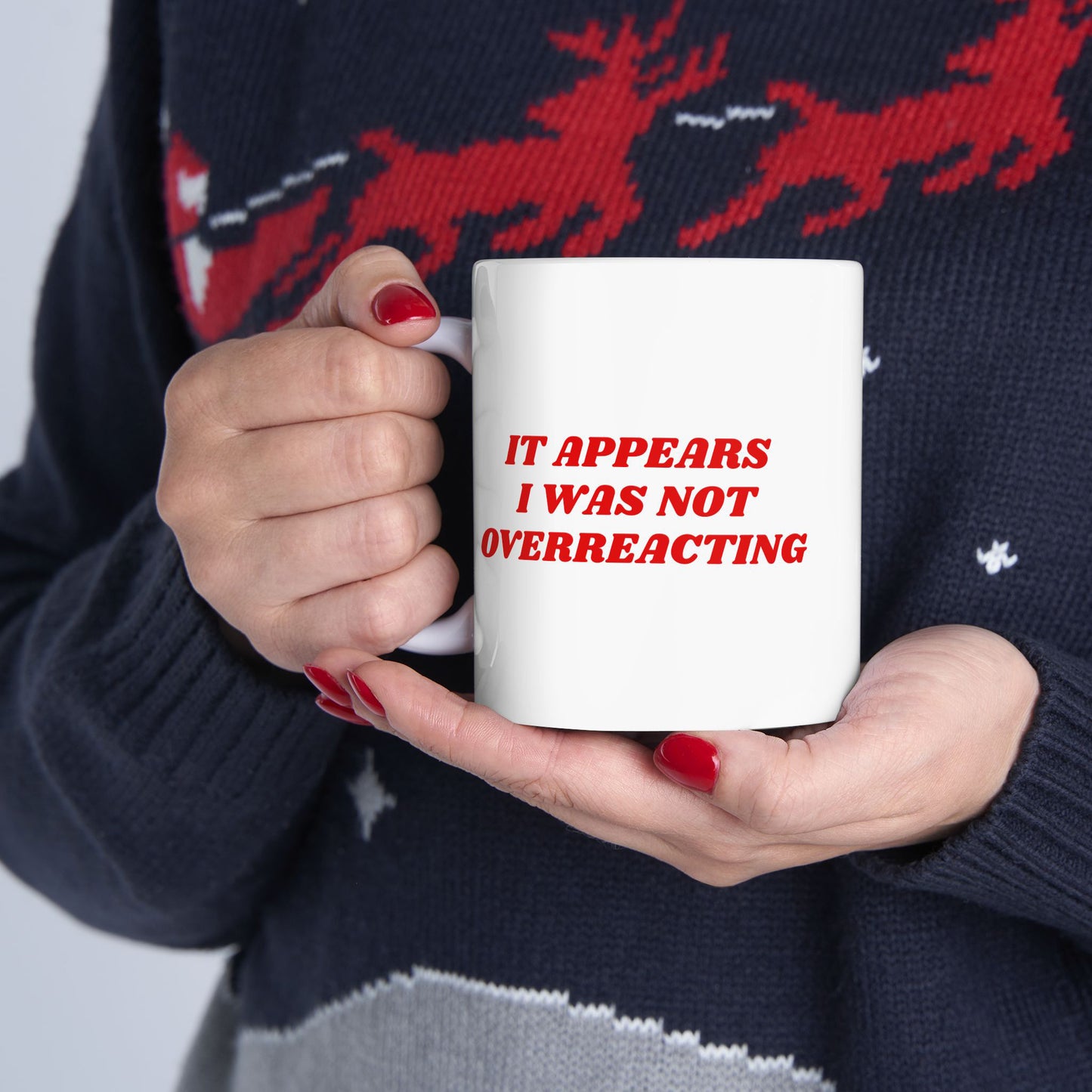 Person holding an Overreacting Mug with the phrase “IT APPEARS I WAS NOT OVERREACTING” in bold red letters, wearing a dark sweater with a red reindeer motif and bright red nail polish.