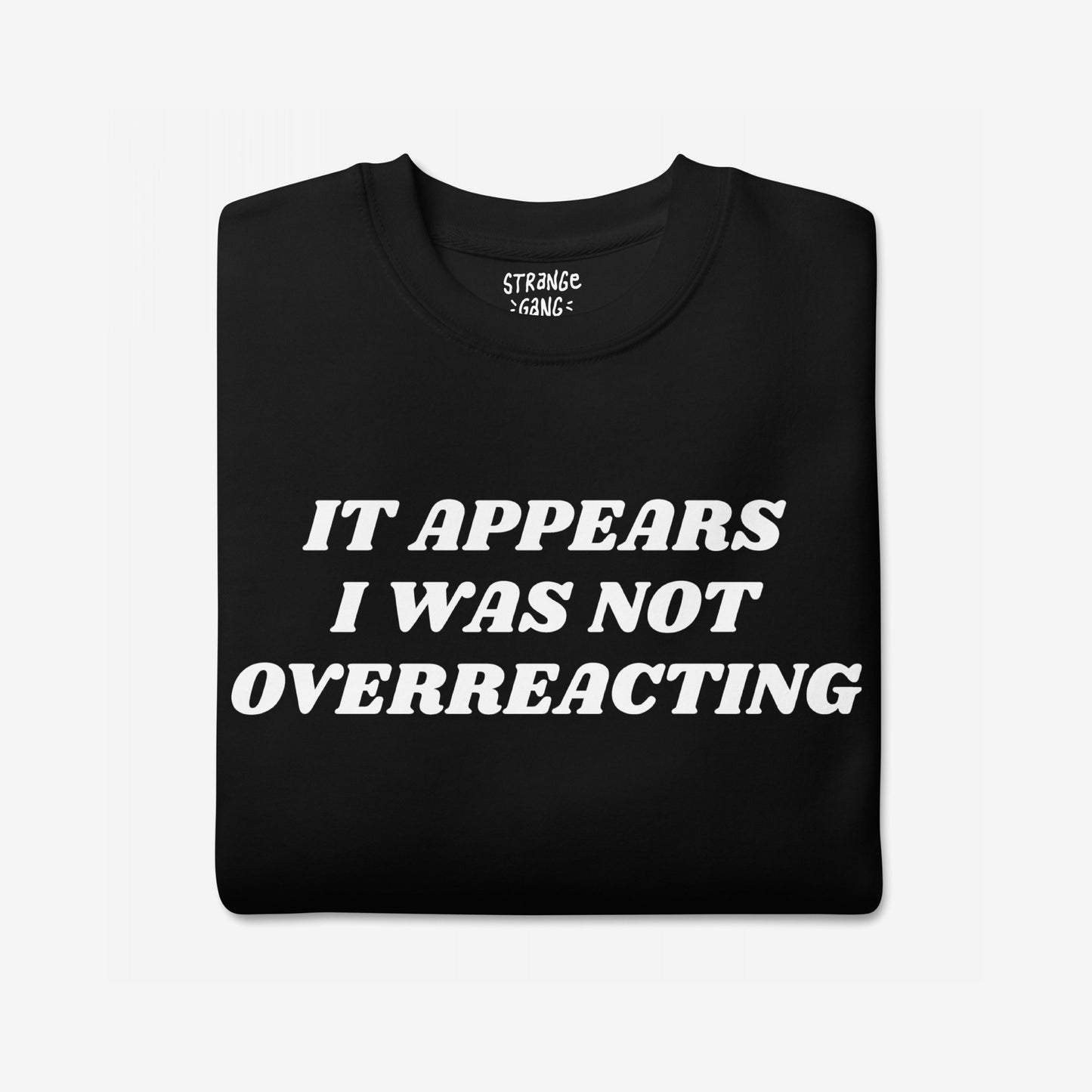 Overreacting Hoodie & Sweatshirt features bold white lettering, “IT APPEARS I WAS NOT OVERREACTING,” on a folded black T-shirt, representing defiance against oppressive systems and advocating for societal resistance.