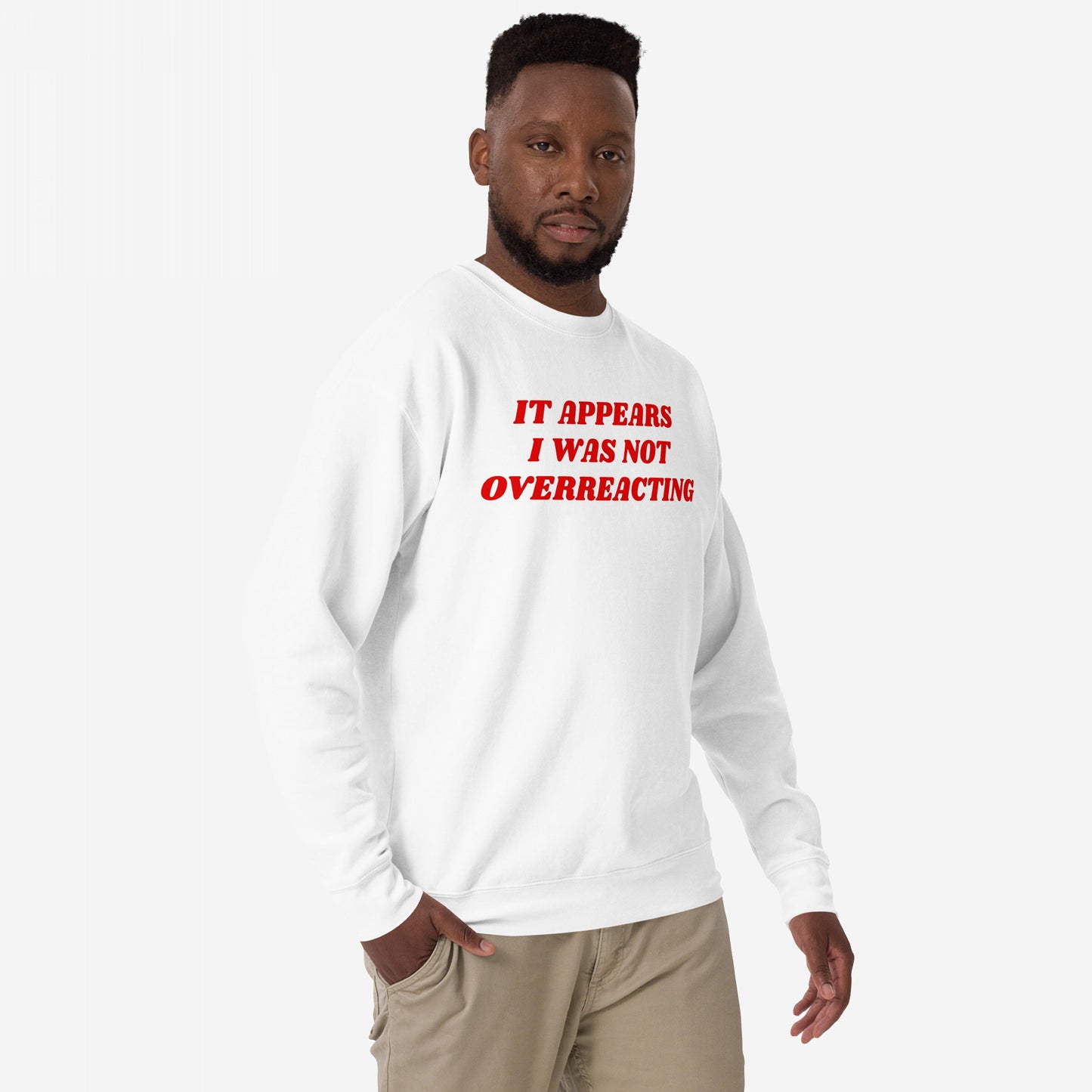Man wearing the Overreacting Hoodie & Sweatshirt with bold red text, embodying resistance and critique against oppressive systems, stands against a plain background, hand in pocket, looking forward.