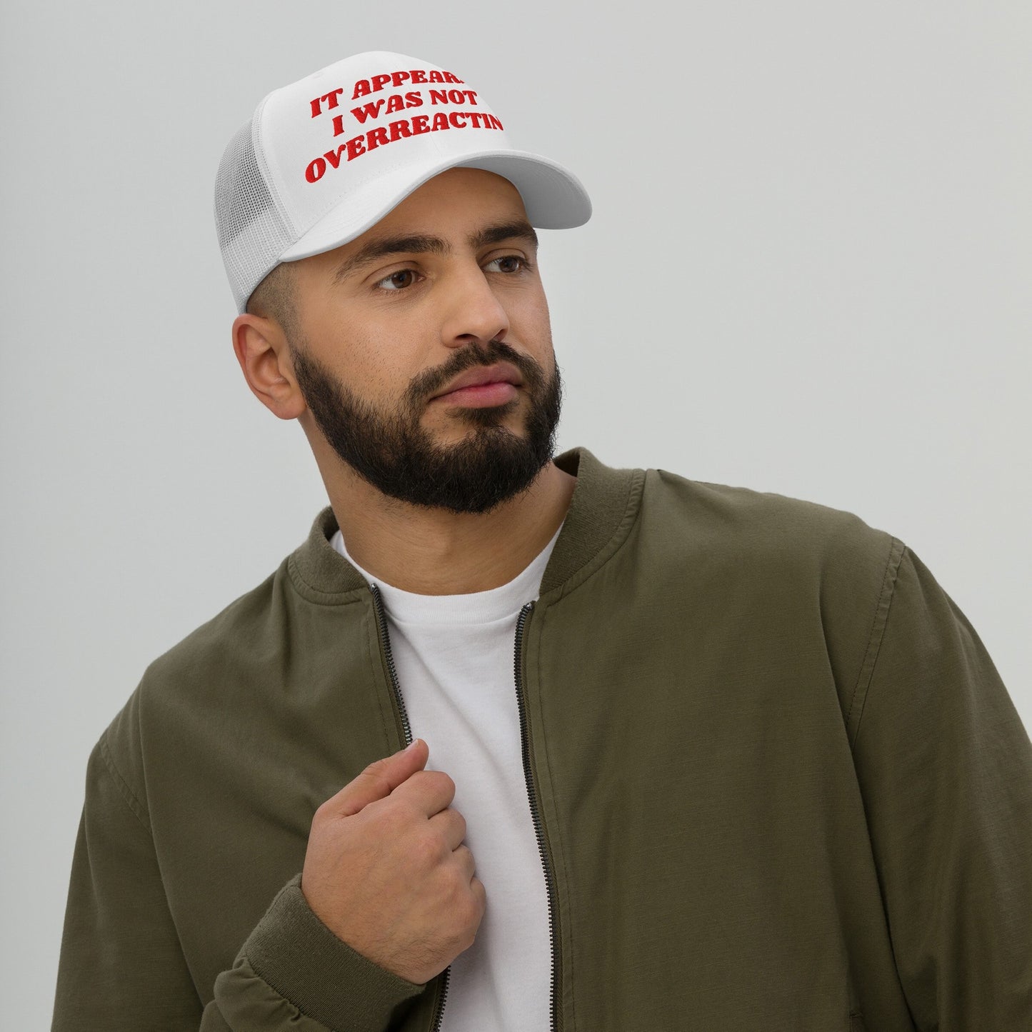 A young bearded man models the Overreacting Hat, a white mesh‐back trucker cap with bold red lettering, expressing defiance against political gaslighting while capturing a progressive spirit.