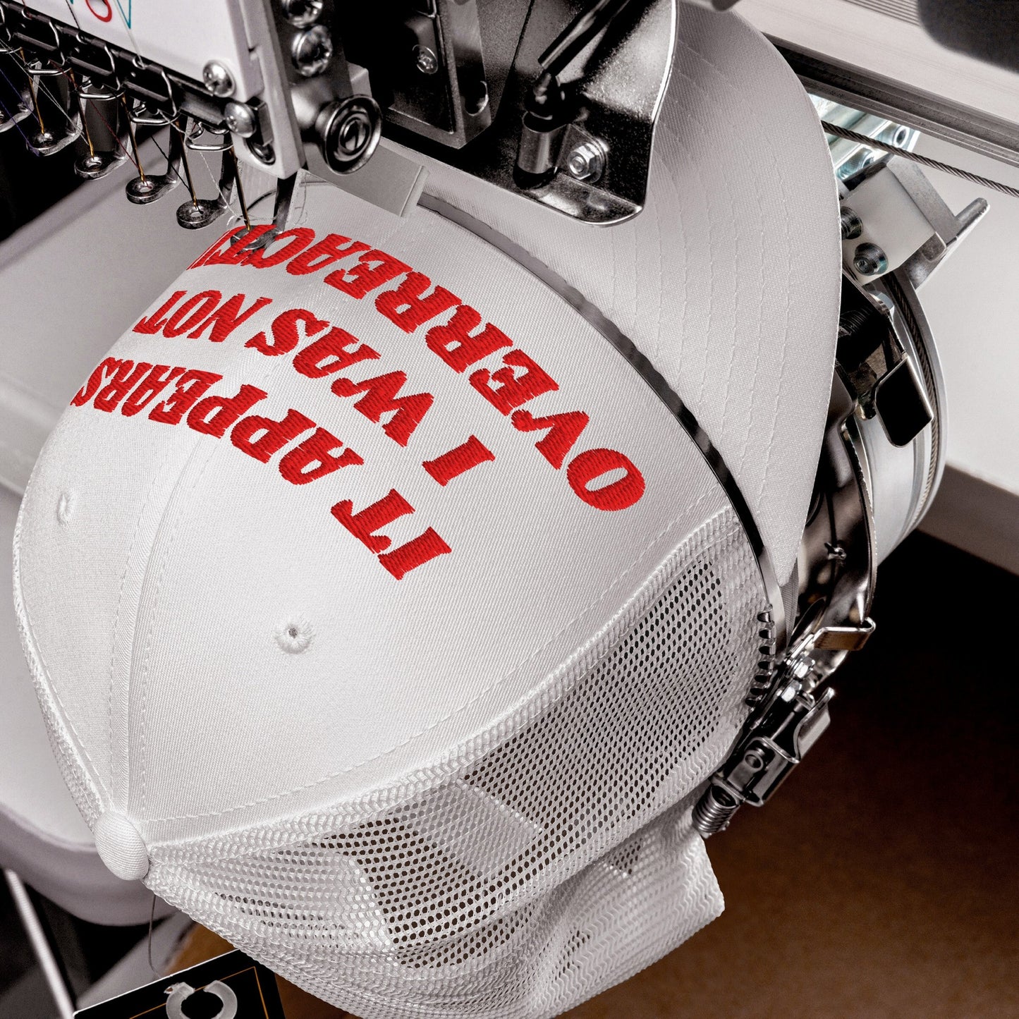 The Overreacting Hat, a white mesh-back cap, is secured in an embroidery machine, stitching bold red letters that confront political gaslighting, embodying activism and defiance against oppressive narratives.