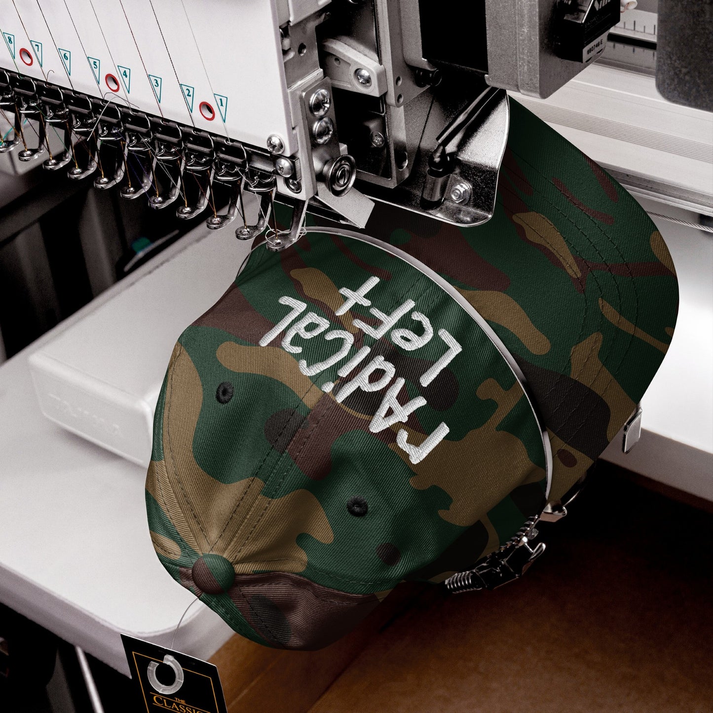 Camo-print baseball cap in embroidery machine, stitching Radical Left on front. Hang tag reads “CLASSIC.” Part of a collection promoting equality and progress.