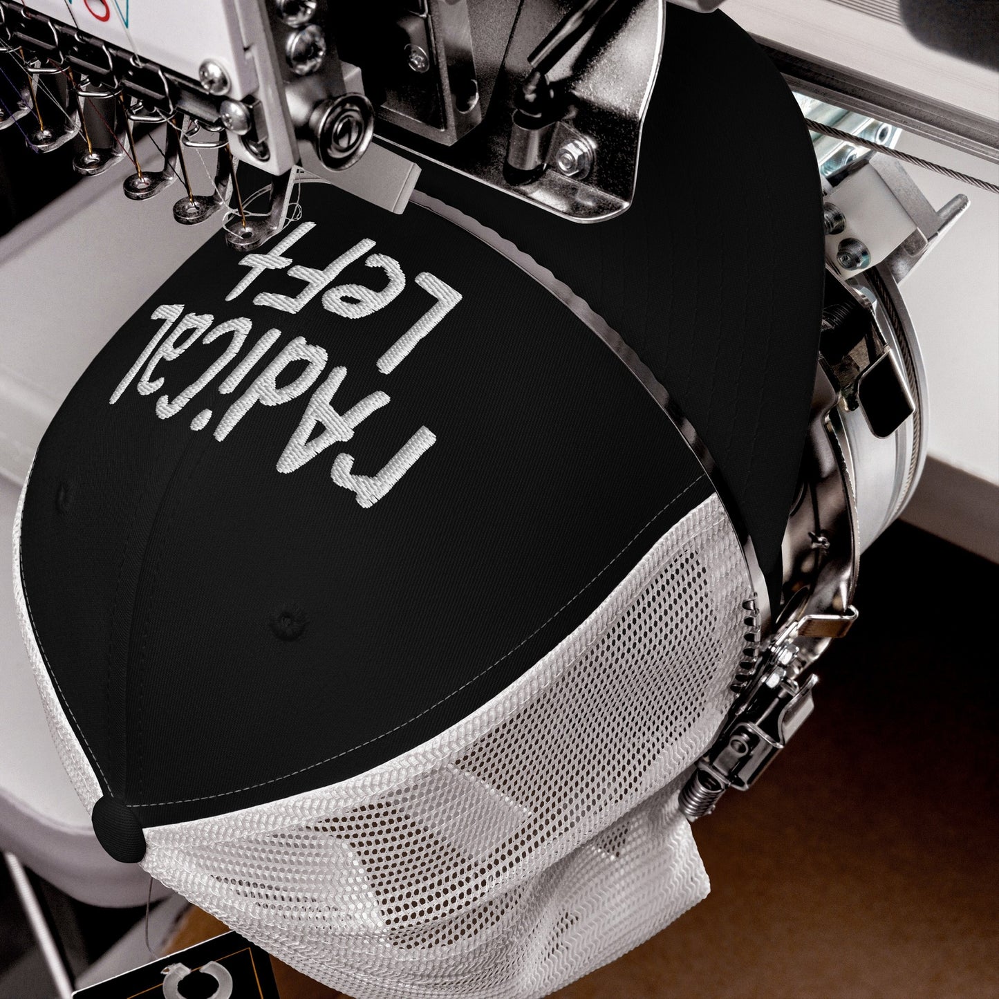 Close-up of an industrial embroidery machine stitching Radical Left in white thread on a black mesh-back trucker hat, symbolizing bold progressivism and a stand against fearmongering.