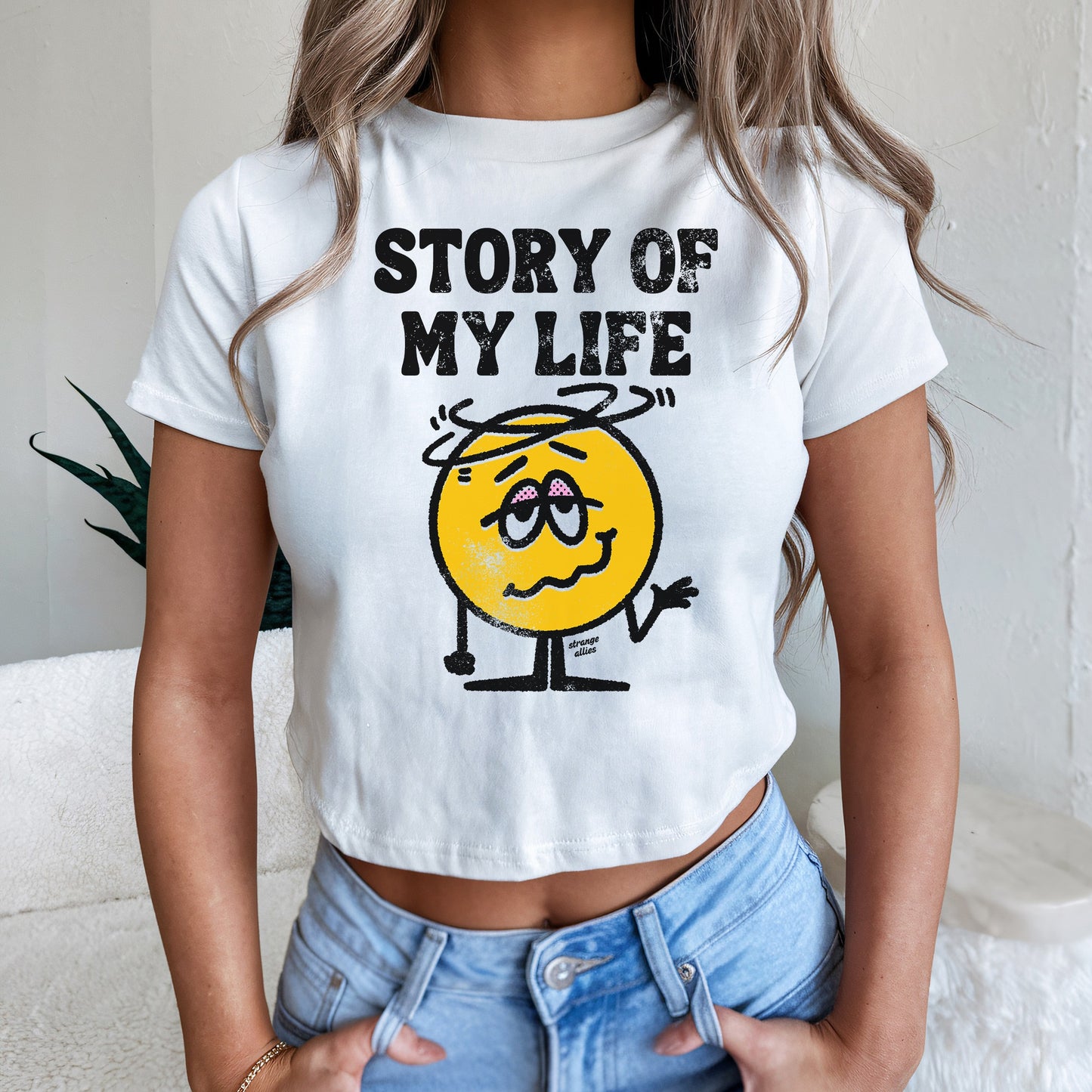 Story Of My Life Women's Baby Tee featuring a dizzy cartoon face and bold lettering, capturing a playful, overwhelmed vibe. The tee is slightly cropped, paired with light-wash jeans.