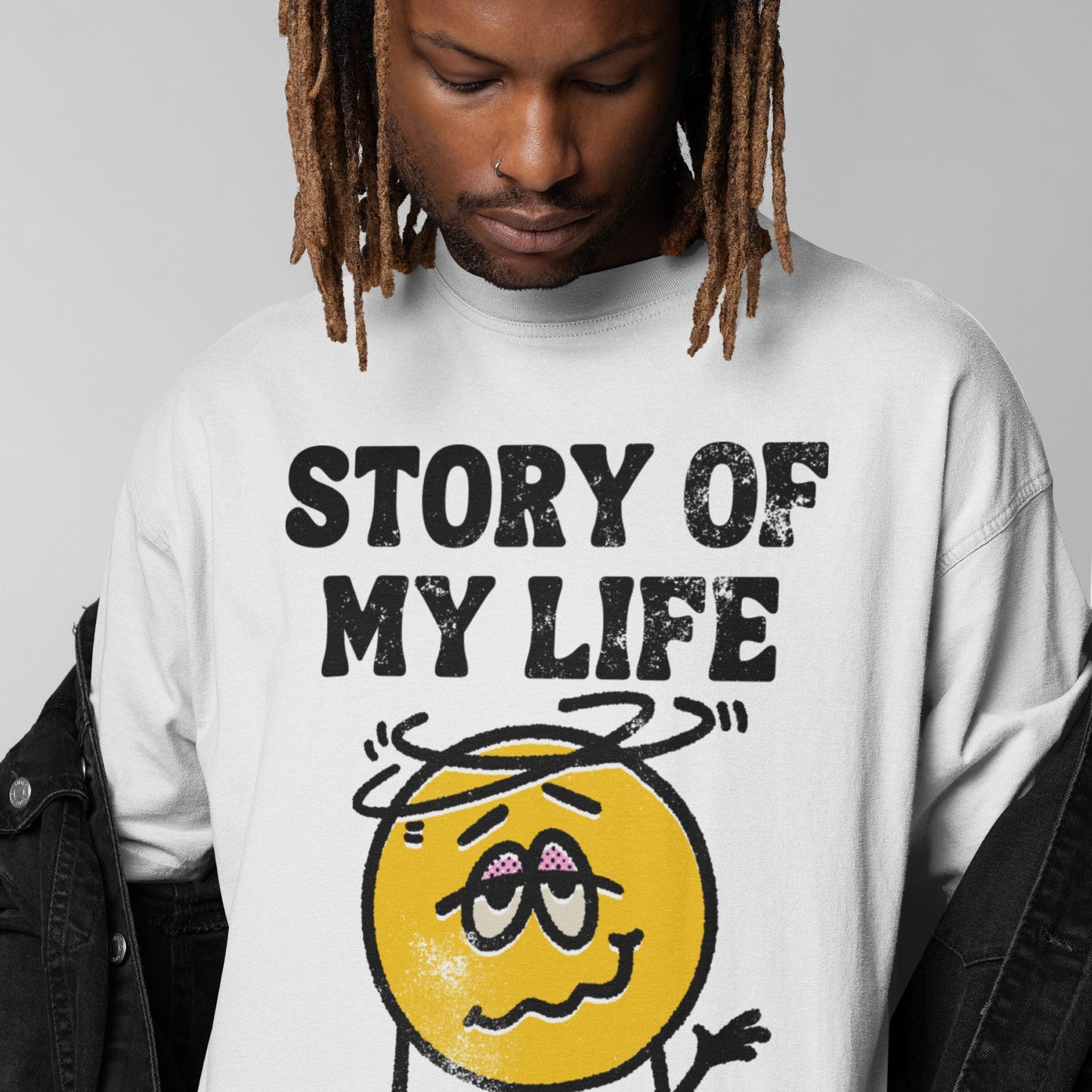 Person wearing a Story Of My Life T-shirt with a distressed slogan and cartoonish dizzy emoticon, styled with dreadlocks and a black jacket, embodying comedic defeat and resilience.