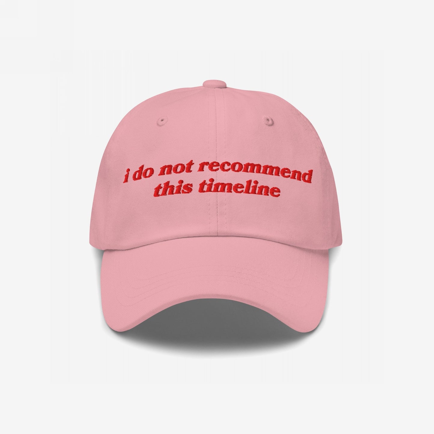 Soft pink Timeline Hat with red vintage-style embroidery stating, “i do not recommend this timeline.” Ideal for expressing political fatigue with style. Available in various styles, supporting Amnesty International.