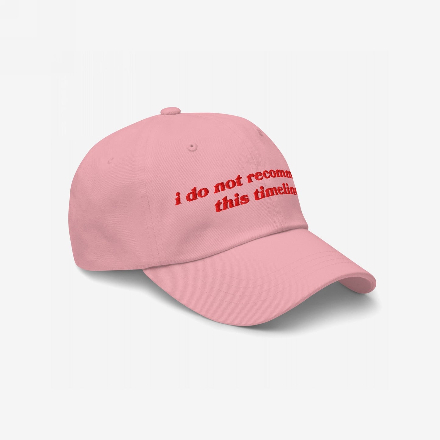 The Timeline Hat features bold red embroidery stating i do not recommend this timeline on a soft pink dad hat, capturing a sarcastic stance on current events with vintage-inspired lettering.