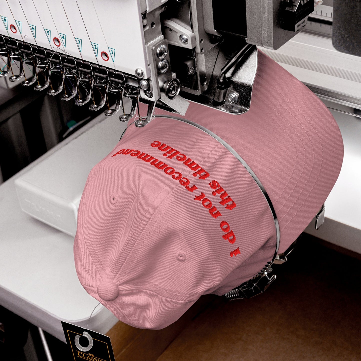 The Timeline Hat in production: An industrial embroidery machine stitches I do not recommend this timeline onto a pink cap, highlighting its sarcastic, politically conscious message.