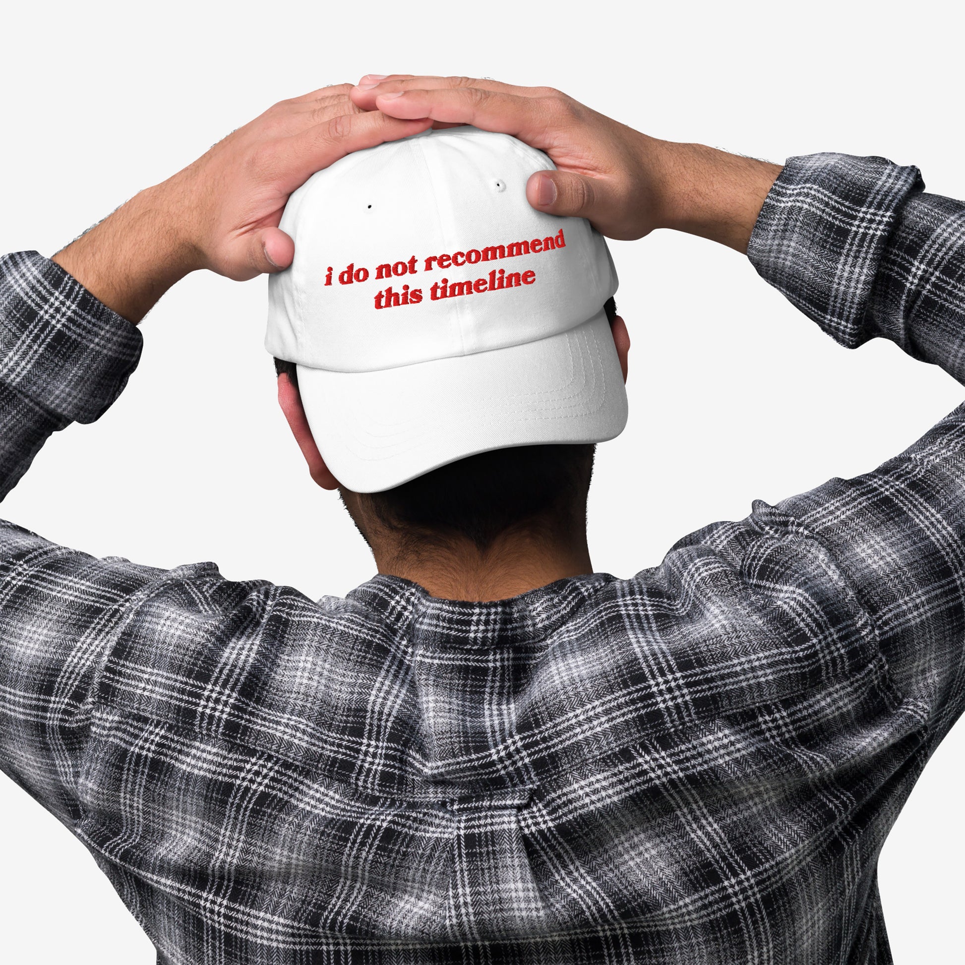 Person wearing the Timeline Hat, a white cap embroidered with “i do not recommend this timeline” in red, paired with a black-and-white plaid shirt, hands resting on the hat.