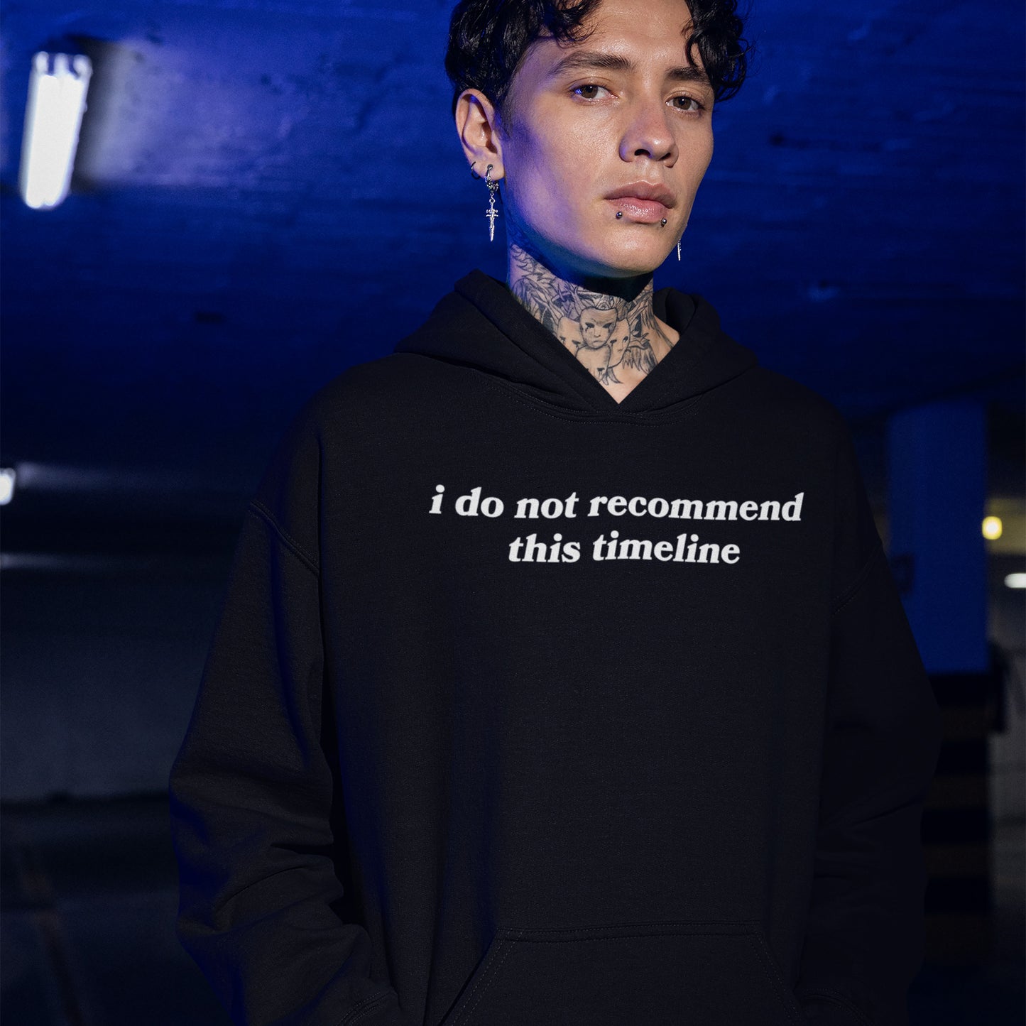 A young person in a dim parking garage wears a black Timeline Shirt hoodie, showcasing neck tattoos and piercings. The shirt humorously critiques modern life, aligning with a bold, expressive style.
