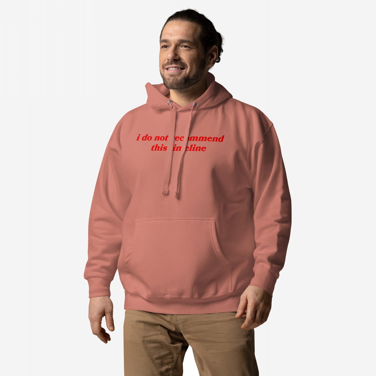 Man wearing the Timeline Hoodie, featuring i do not recommend this incline in red serif text, smiles subtly. The muted coral-pink hoodie pairs with tan pants, embodying sarcastic dystopian style.