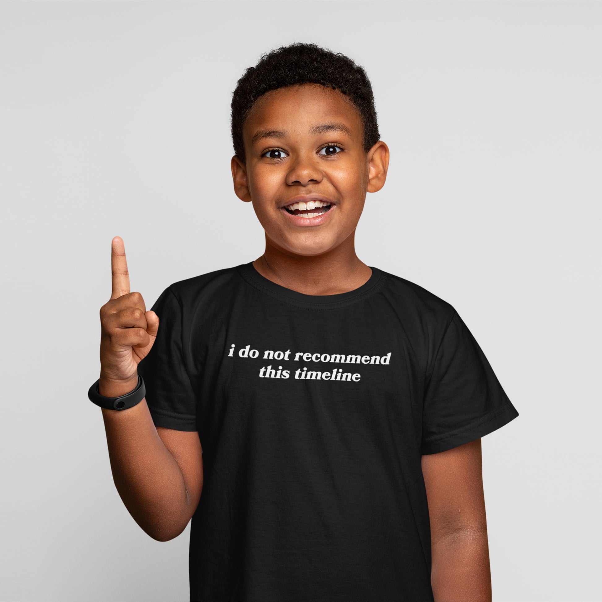 Young boy animatedly holds up one finger, wearing the Timeline Shirt with the phrase “i do not recommend this timeline,” capturing humor and critique. Black wristband on left wrist.