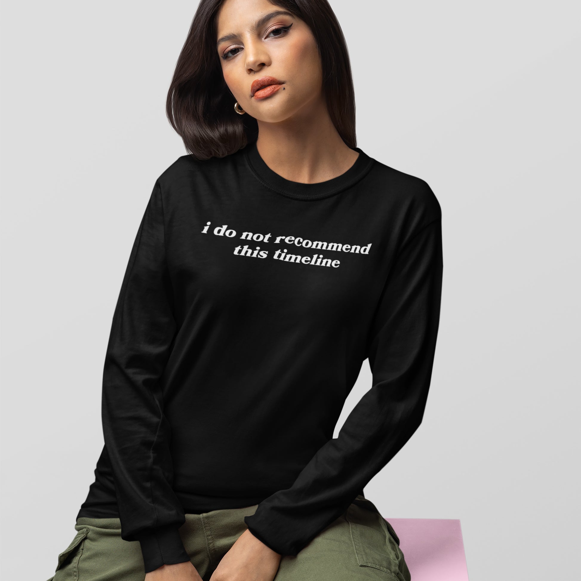 Young woman in black long-sleeve Timeline Shirt with white text, paired with olive cargo pants, seated on a pink block, embodying humor and critique for today's world.