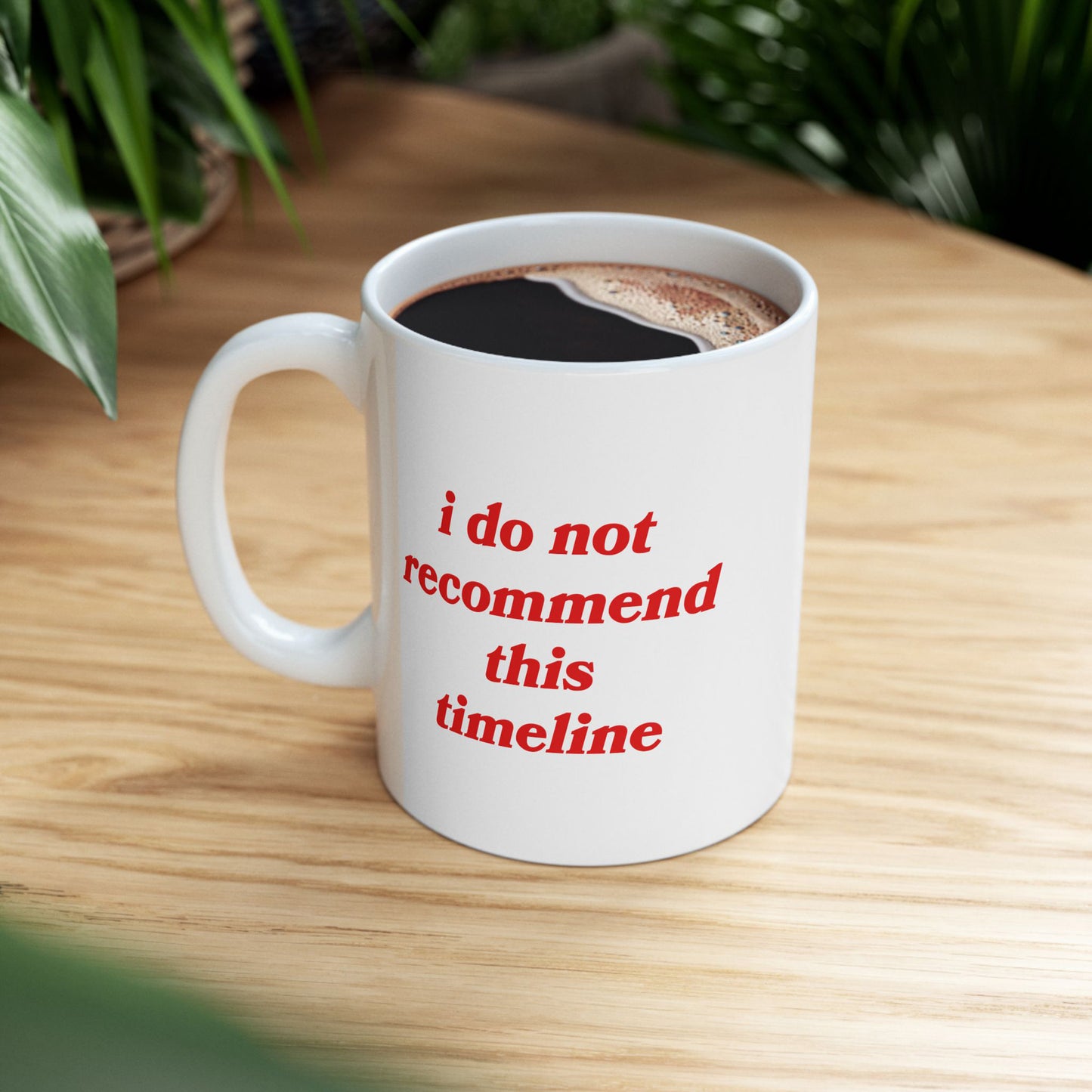 The Timeline Mug, a white ceramic cup with bold red text, humorously critiques societal chaos, filled with black coffee, set on a wooden table amidst leafy plants, reflecting a cozy, introspective vibe.