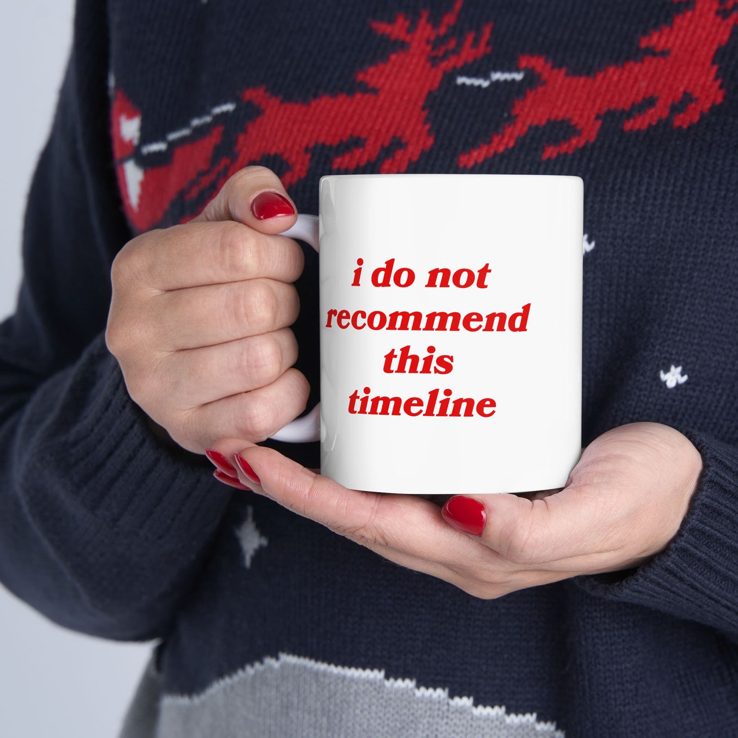 Hands with red-painted nails hold the Timeline Mug, featuring the phrase “i do not recommend this timeline” in red, against a dark blue sweater with a red reindeer pattern, embodying snarky existential commentary.
