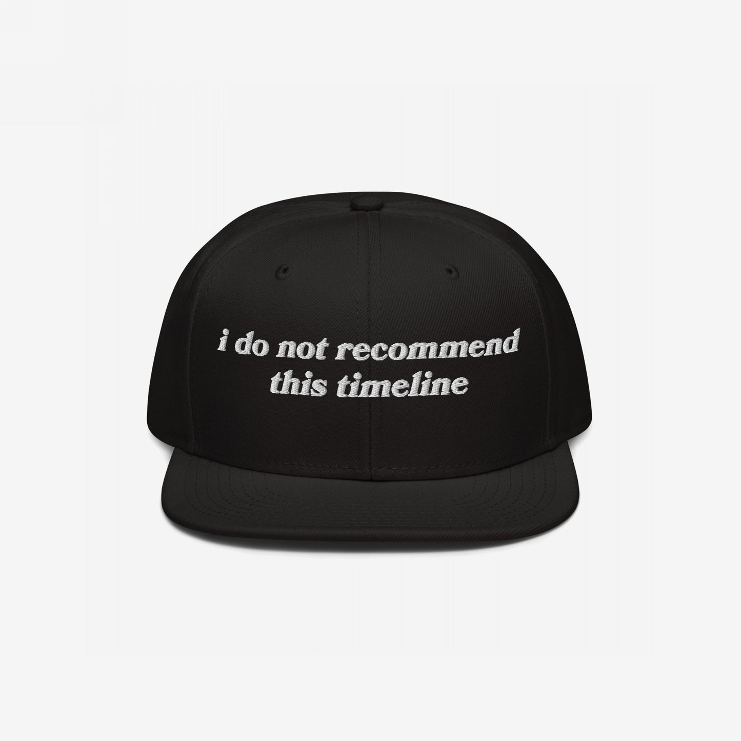 Black Timeline Hat with white embroidery reading, “I do not recommend this timeline.” Ideal for expressing political fatigue and sarcasm. Soft snap-back design against a plain backdrop. Ships from the E.U.