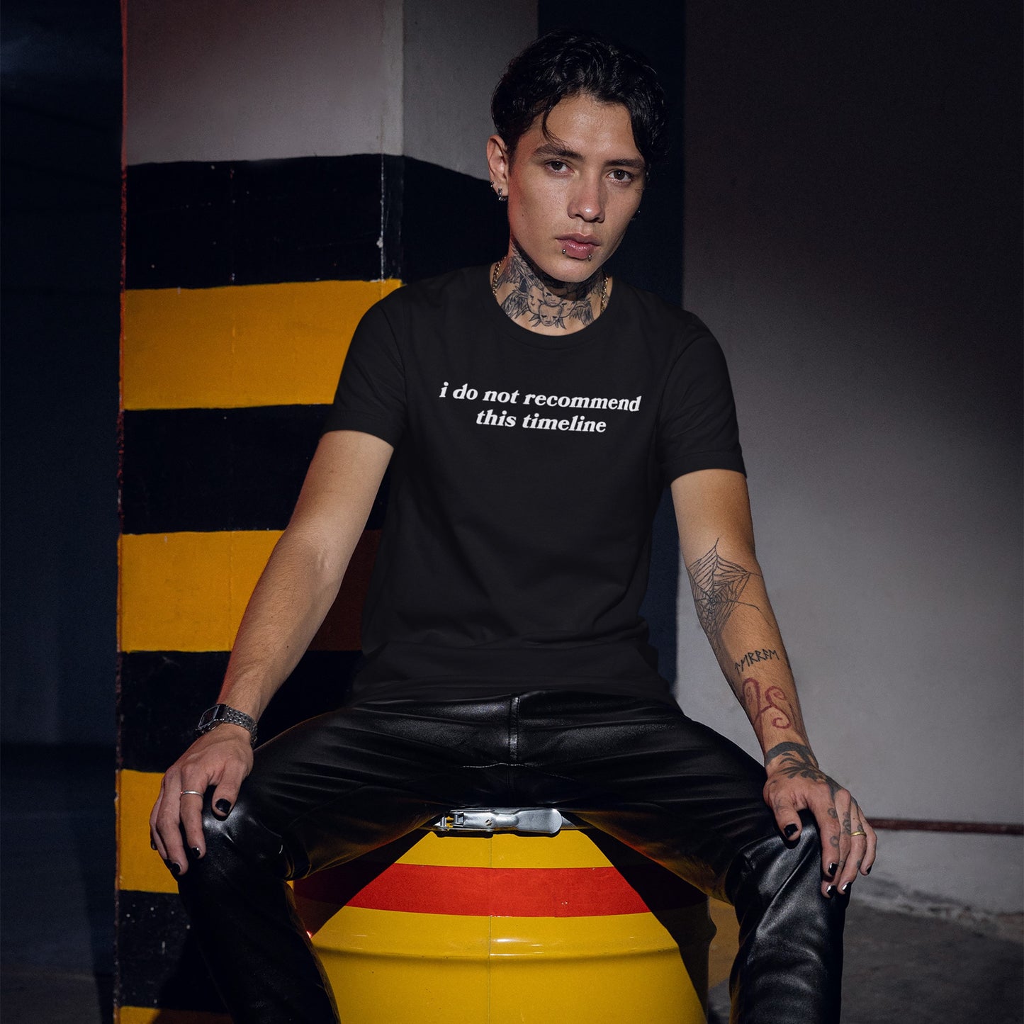 Young man in a Timeline T-shirt with the phrase “i do not recommend this timeline,” sitting on a yellow drum in a moody industrial setting, emphasizing edgy style with tattoos and piercings.