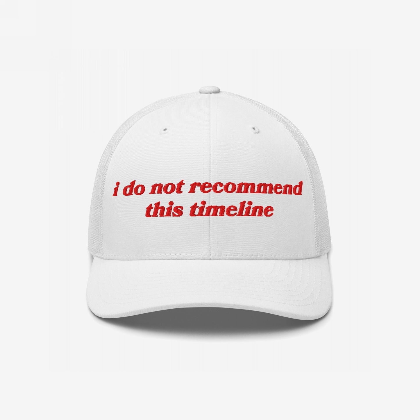 White trucker-style baseball cap with red embroidery reading “I Do Not Recommend This Timeline,” signaling political discontent. Reflects sarcastic, protest-driven ethos, embodying a soft fit for hard truths.