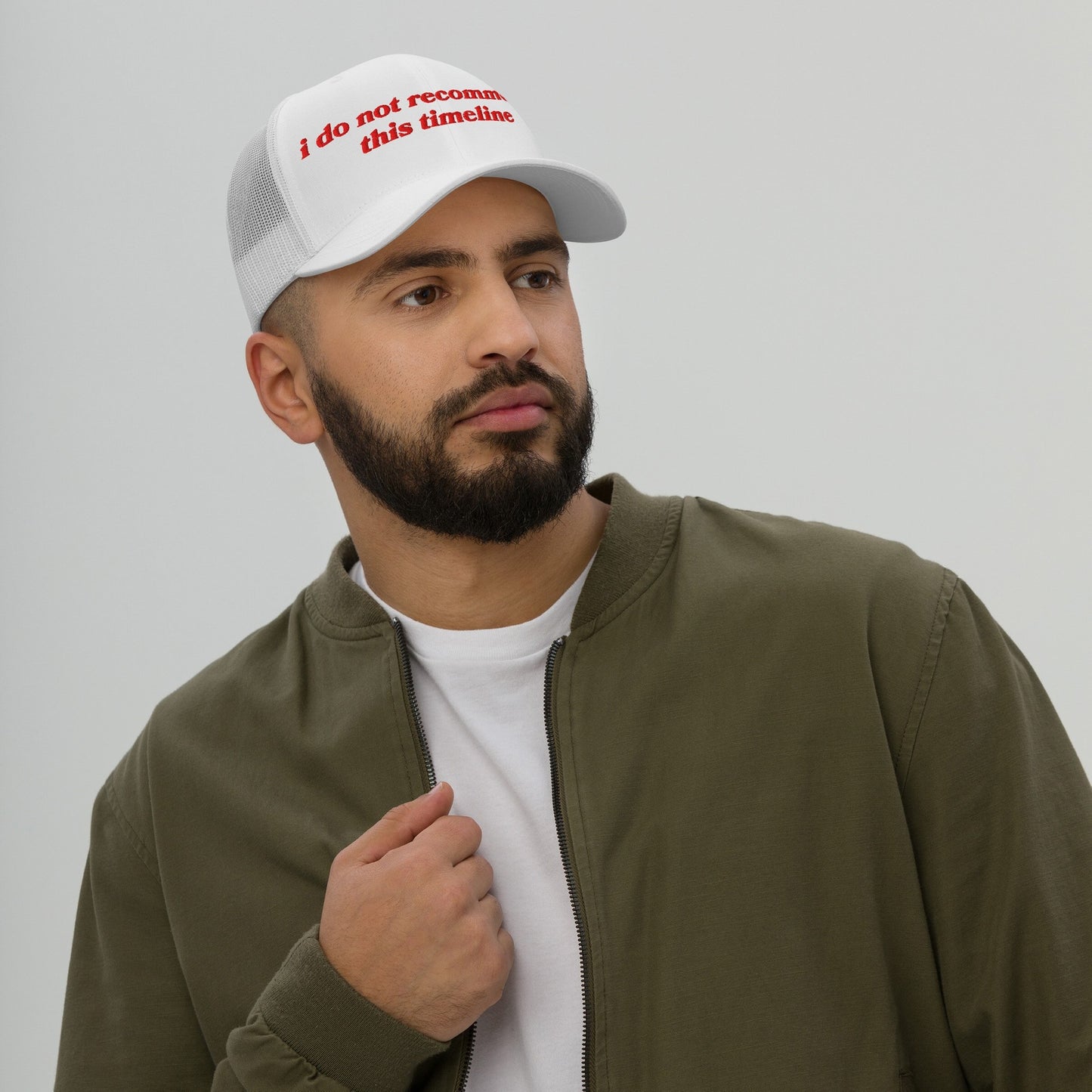 A bearded man wears the Timeline Hat, a white mesh-back trucker cap with “i do not recommend this timeline” in red, paired with a white T-shirt and olive-green jacket.