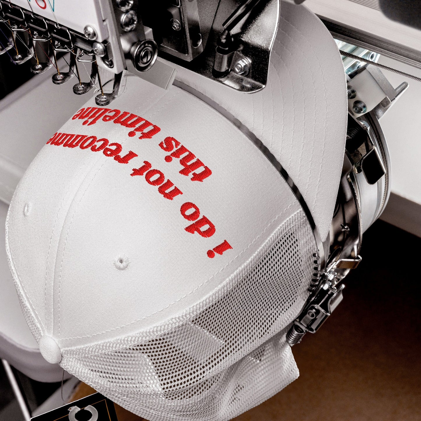 White mesh-back baseball cap in embroidery machine, featuring red text I do not recommend this timeline, capturing sarcastic political sentiment. Needles poised above, actively stitching the Timeline Hat's design.