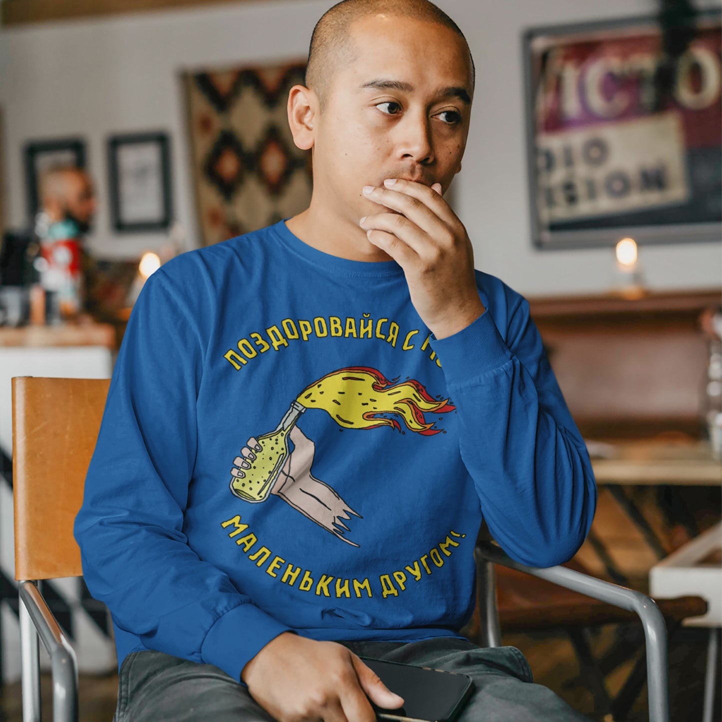Man seated wearing the Say Hello Ukraine T-shirt, featuring a hand-drawn Molotov cocktail and bold Russian text, symbolizing resistance and defiance. He holds a smartphone and appears contemplative in a café setting.