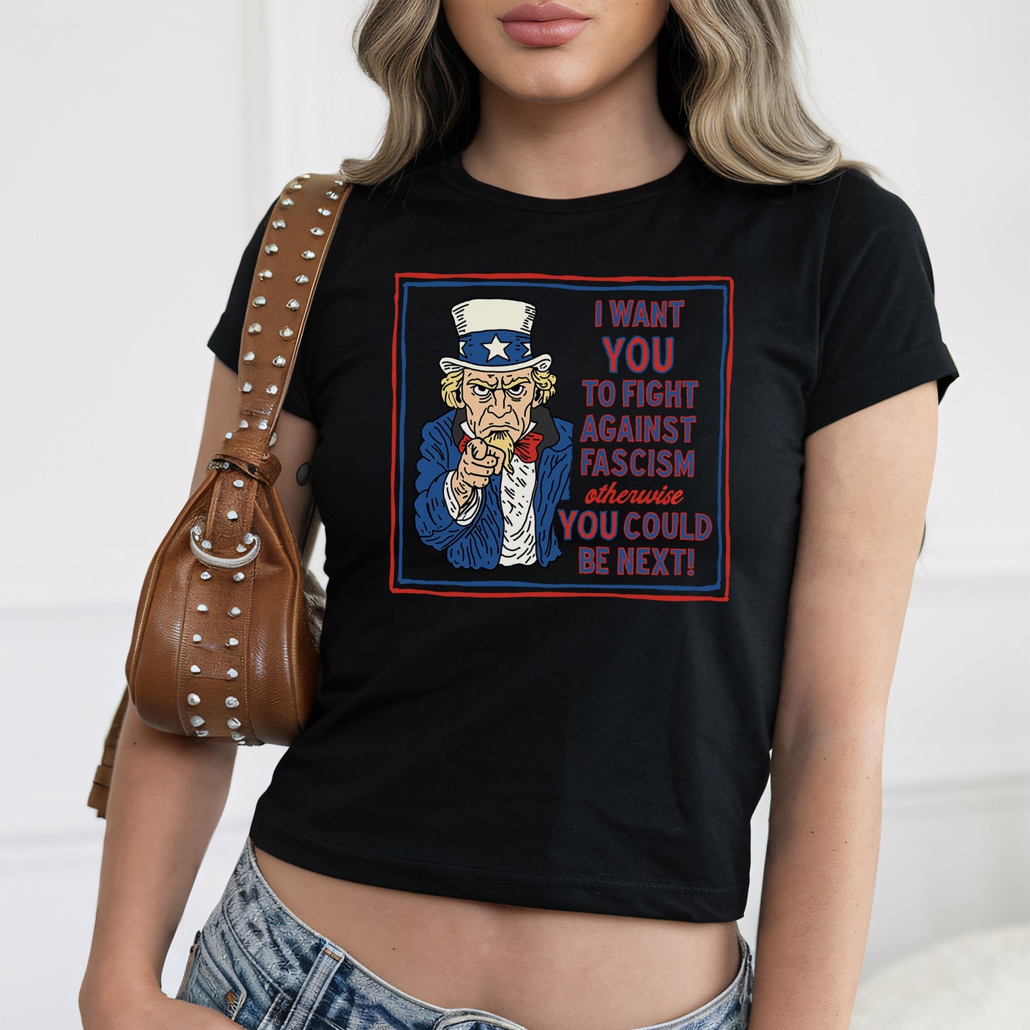 Person wearing Anti-Fascist Uncle Sam Women's Baby Tee with stylized Uncle Sam graphic and bold anti-fascism slogan, paired with studded brown shoulder bag and low-rise blue jeans.