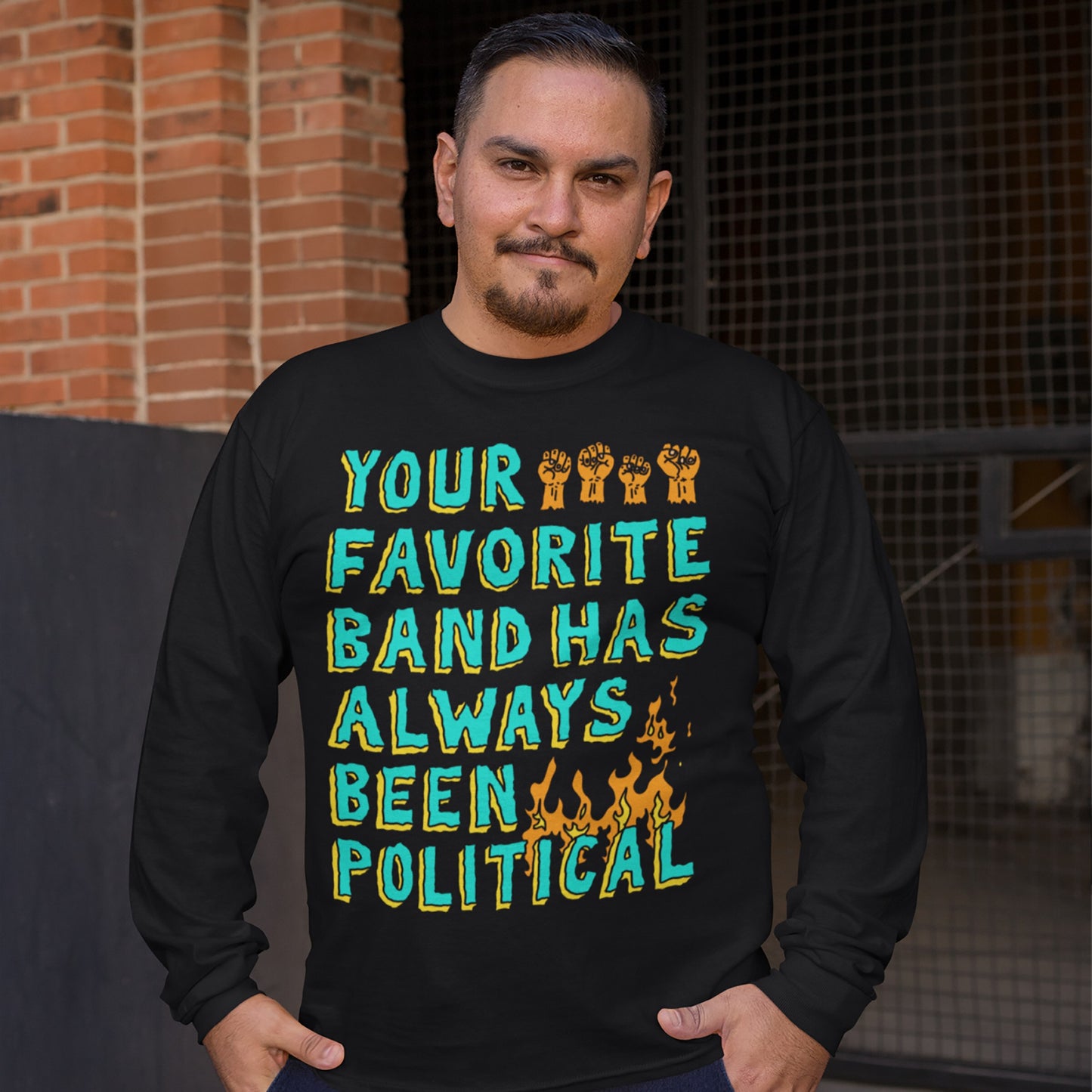 Man outdoors wearing Your Favorite Band Has Always Been Political long-sleeve T-shirt, featuring bold lettering, stylized fists, and flames. Reflects protest anthem energy and art as resistance.
