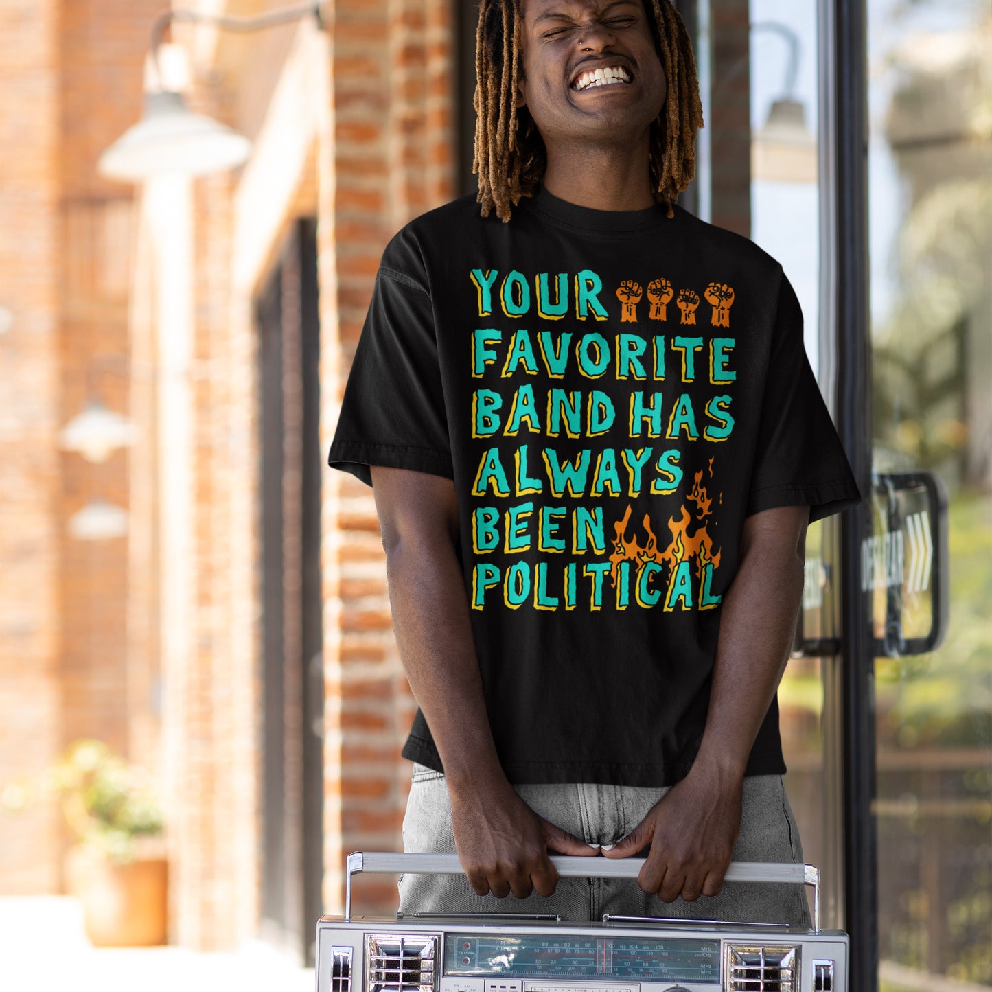 Young person grinning, holding a boombox, wears a Your Favorite Band Has Always Been Political black T-shirt. The design includes raised fists and flames, embodying punk-inspired, rebellious spirit.