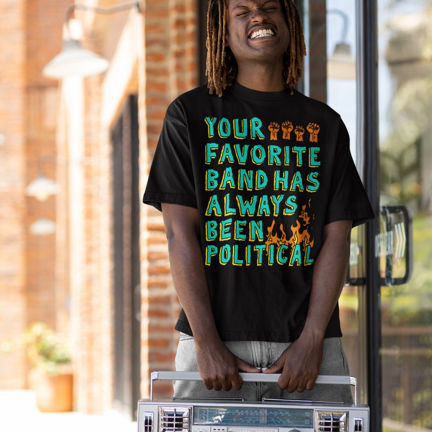 A young person grinning broadly outdoors, wearing the Your Favorite Band Has Always Been Political T-shirt featuring bold lettering, raised-fist icons, and flames, while holding a vintage silver boombox.