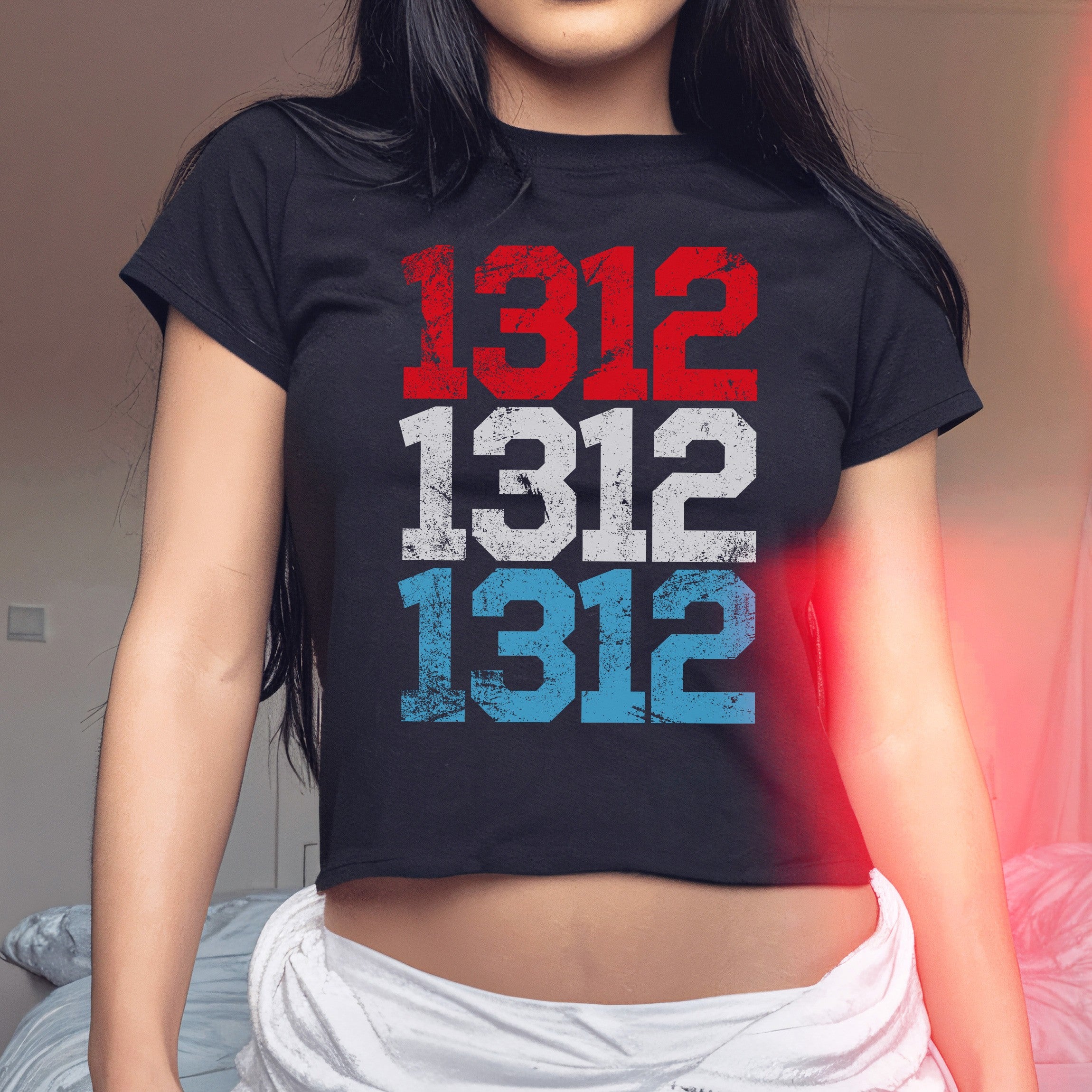 A person models the 1312 ACAB Women's Baby Tee, featuring the number 1312 stenciled thrice in red, white, and blue, paired with light-colored, loose bottoms, embodying a protest-ready stance.