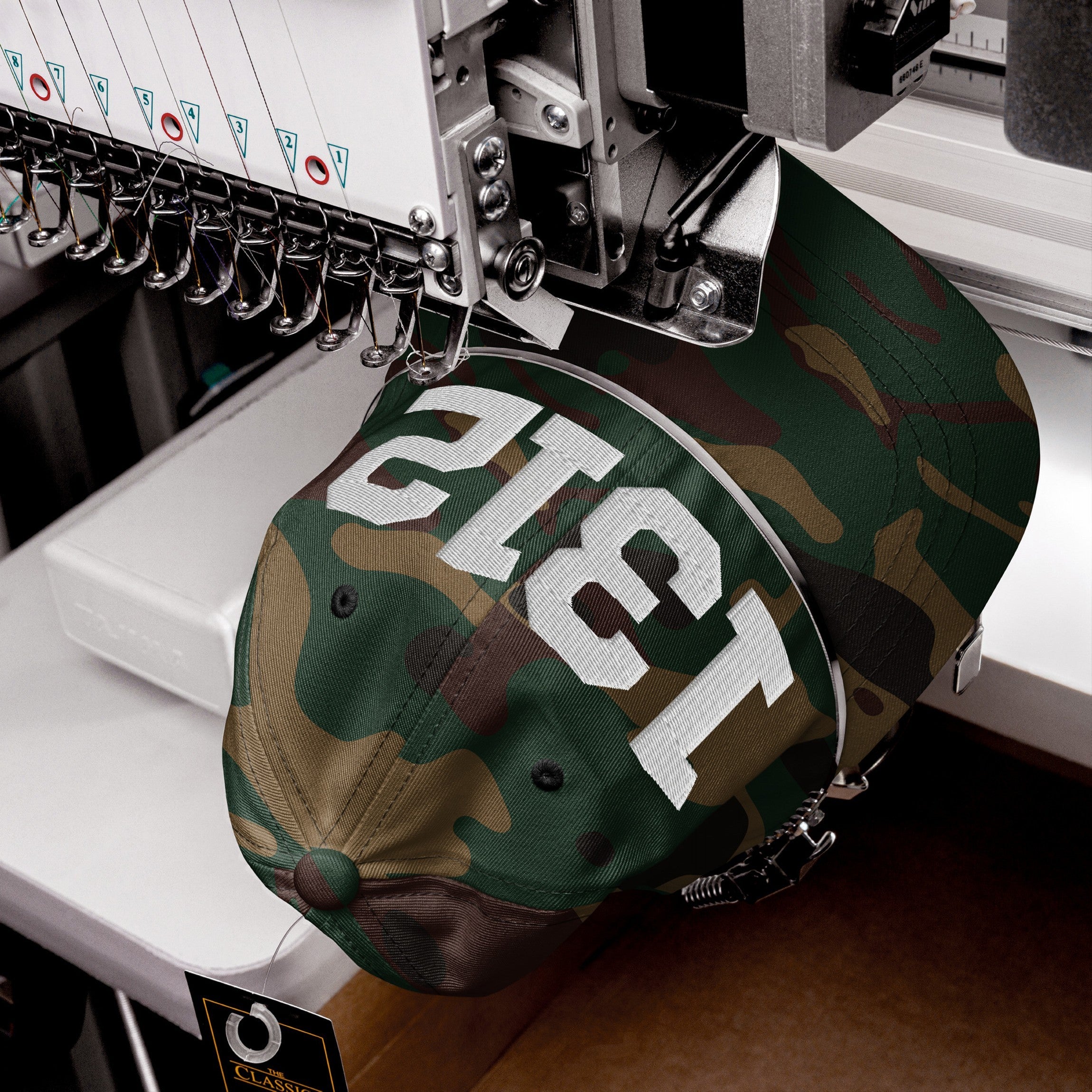 1312 ACAB Hat in camouflage print is being embroidered with white block numbers on an industrial machine, emphasizing protest and accountability. A Classic tag hangs from the brim.