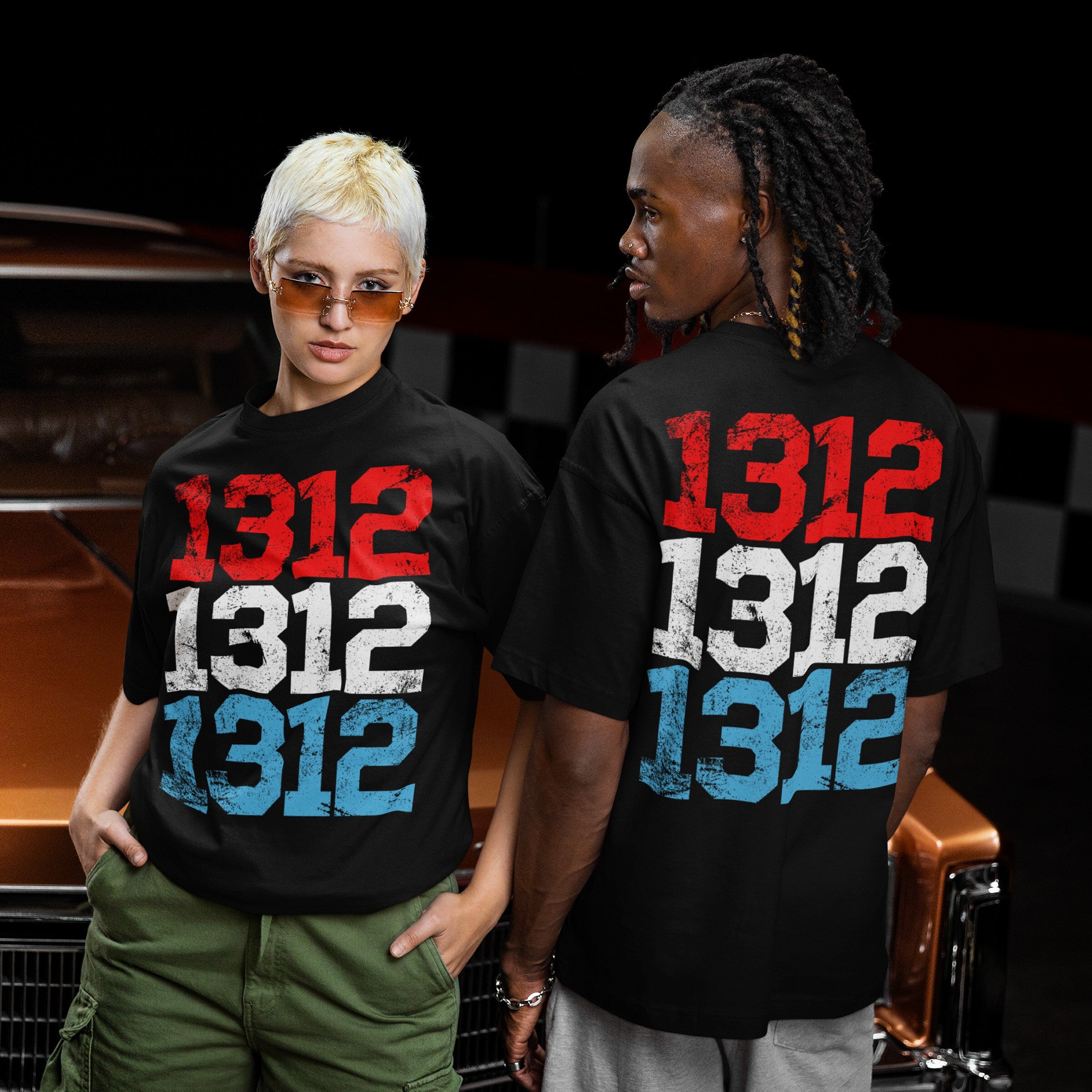Two young adults showcase the 1312 ACAB T-shirt. One faces forward; the other, backward. Both display 1312 in red, white, and blue, embodying a bold, protest-driven streetwear style.