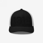 Black-and-white trucker cap with 1312 embroidered in black on the front, symbolizing ACAB. The cap features black front panels and brim, with white mesh sides and back.