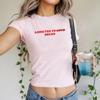 Person wearing the Addicted To Love / Drugs Women's Baby Tee, featuring bold text with LOVE crossed out for DRUGS, paired with denim jeans and holding a blue iced drink.