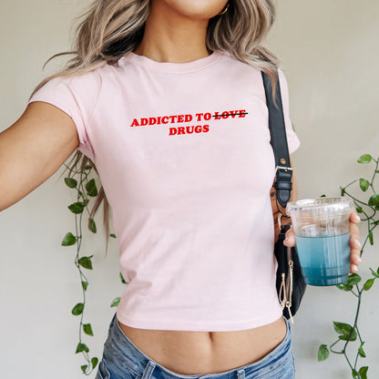 Person wearing the Addicted To Love / Drugs Women's Baby Tee, featuring bold text with LOVE crossed out for DRUGS, paired with denim jeans and holding a blue iced drink.