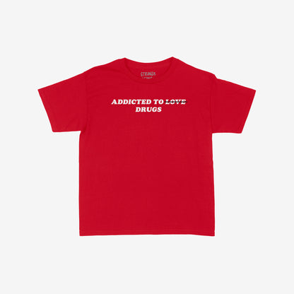 Addicted To Love / Drugs Women's Baby Tee featuring bold white text with LOVE struck-through and replaced by DRUGS, capturing chaotic humor and honesty on a plain red crew-neck style.