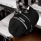 Close-up of a black Addicted To Love / Drugs Hat being embroidered with white and red thread on a sewing machine.