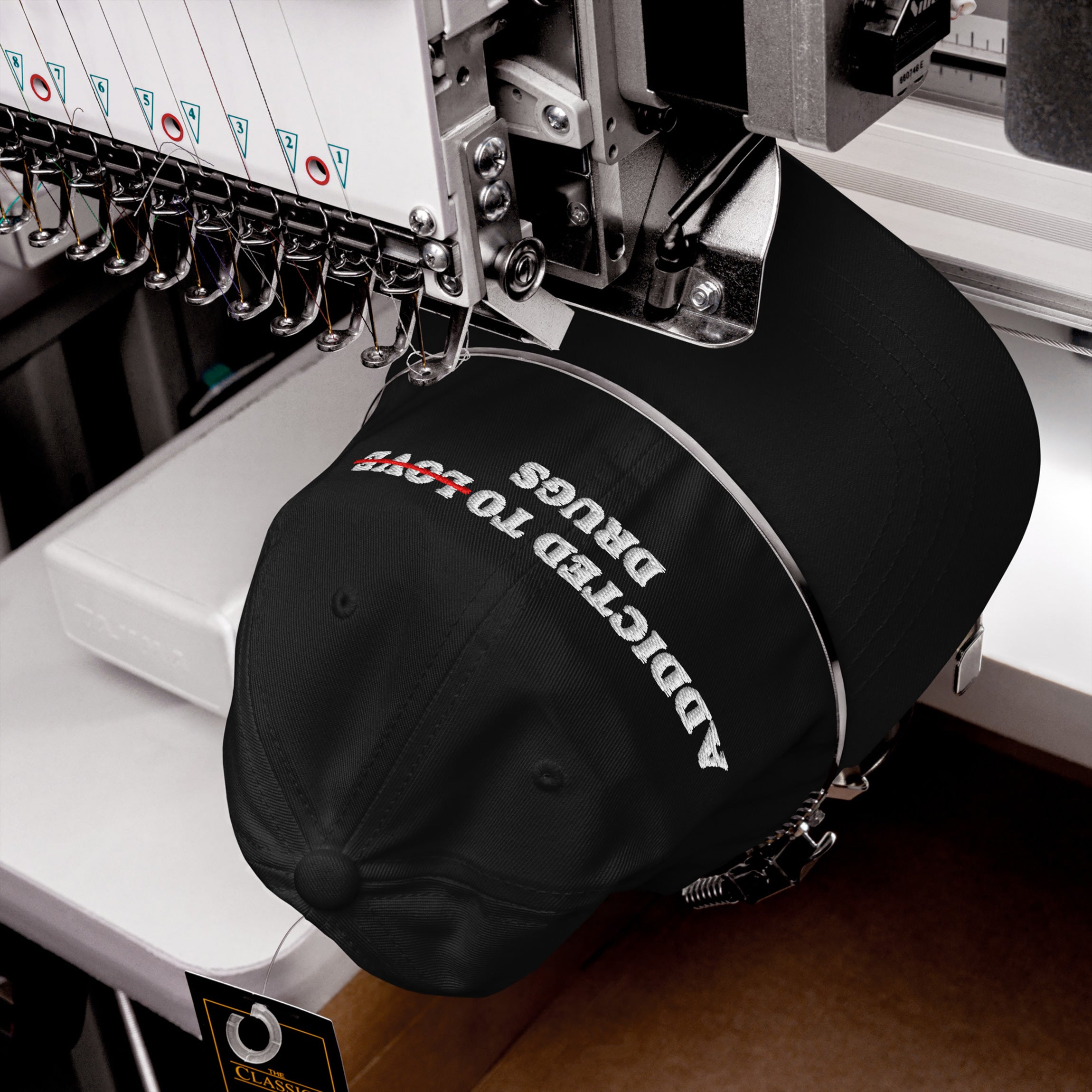 Close-up of a black Addicted To Love / Drugs Hat being embroidered with white and red thread on a sewing machine.