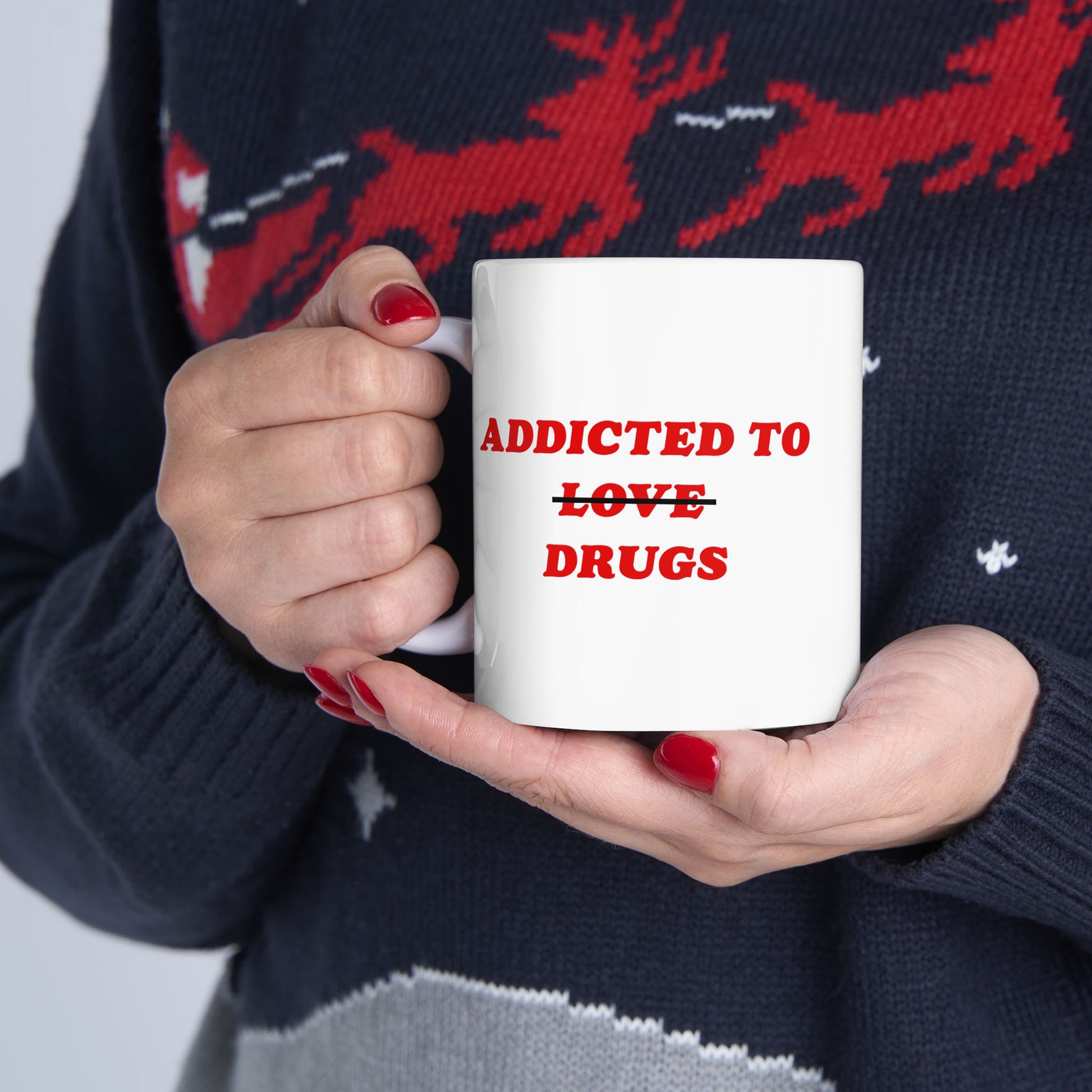 Hands with red nails hold the Addicted To Love / Drugs Mug, featuring bold red text with a humorous strikethrough design. The person wears a dark blue sweater with a red reindeer pattern.