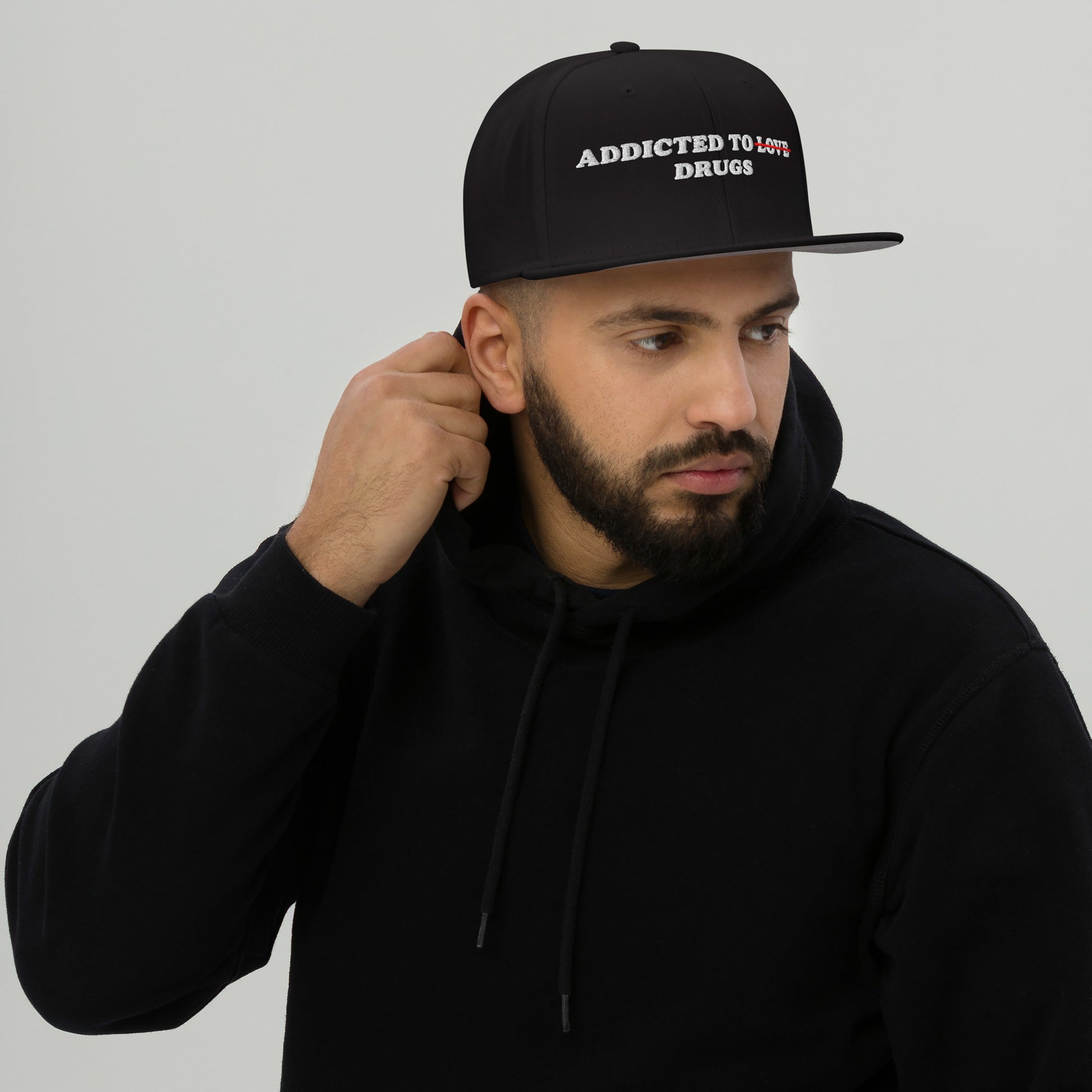 Man wearing a black snapback featuring embroidered text "Addicted To Love" crossed out and replaced by "Drugs" hat titled Addicted To Love / Drugs Hat.