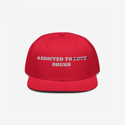 Bright red, flat-brim snapback cap with Addicted To Love/Drugs Hat embroidered. LOVE is struck through, displaying DRUGS beneath, symbolizing raw honesty over romantic illusions.