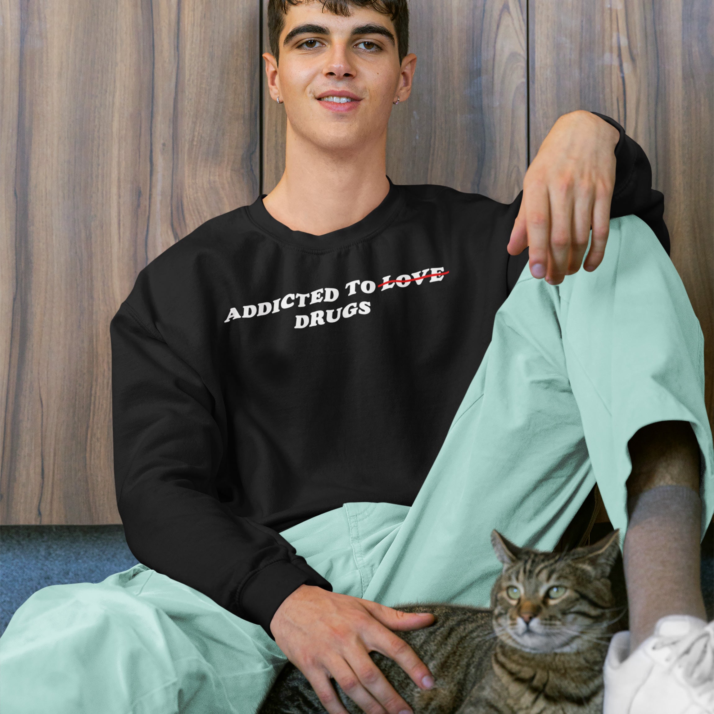 Young man wearing black Addicted To Love / Drugs Shirt with crossed out “Love” replaced by “Drugs,” sitting with cat against wood background
