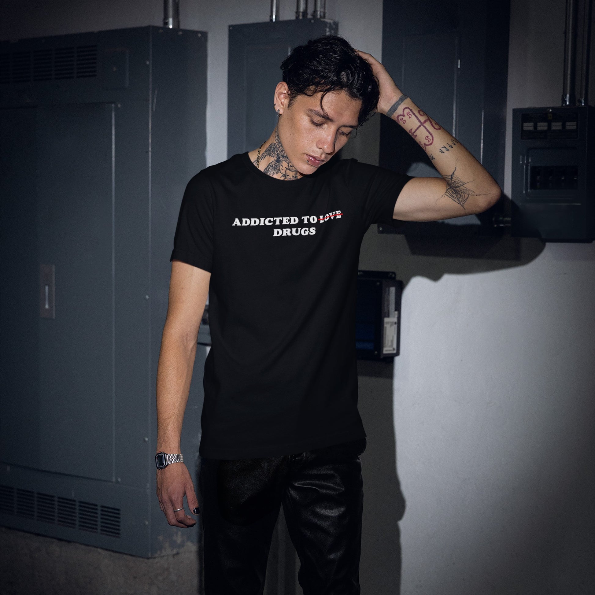 A young person in an industrial setting wears the Addicted To Love / Drugs T-shirt, featuring a bold statement with a crossed-out Love and prominent Drugs, reflecting edgy, unapologetic style.