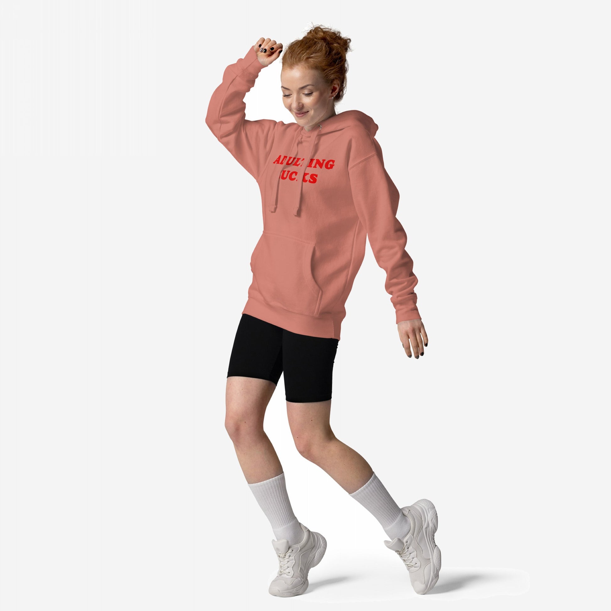 Young woman playfully poses in the Adulting Sucks Hoodie & Sweatshirt, showcasing its bold statement print. She's styled with bike shorts, socks, and sneakers, embodying a carefree, relatable vibe.