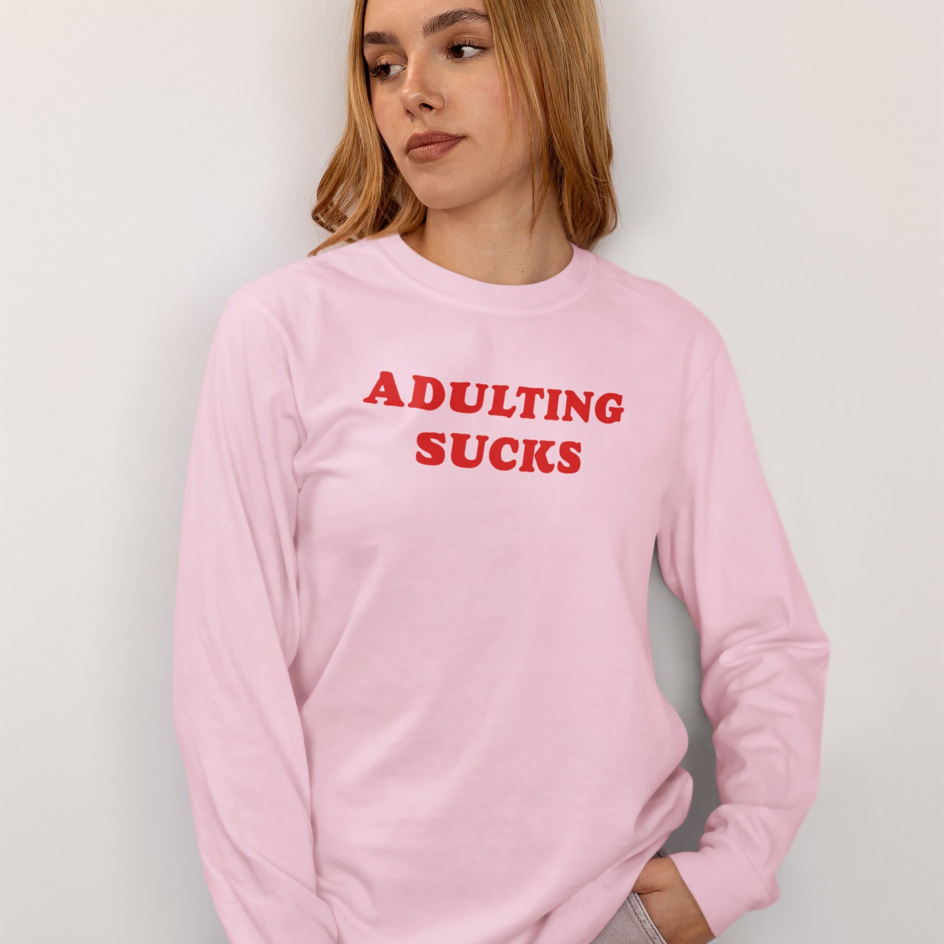 Young person wearing the Adulting Sucks T-shirt, featuring a bold red slogan on a pastel-pink long-sleeve shirt. The design combines retro charm and humor, ideal for expressing modern frustrations.