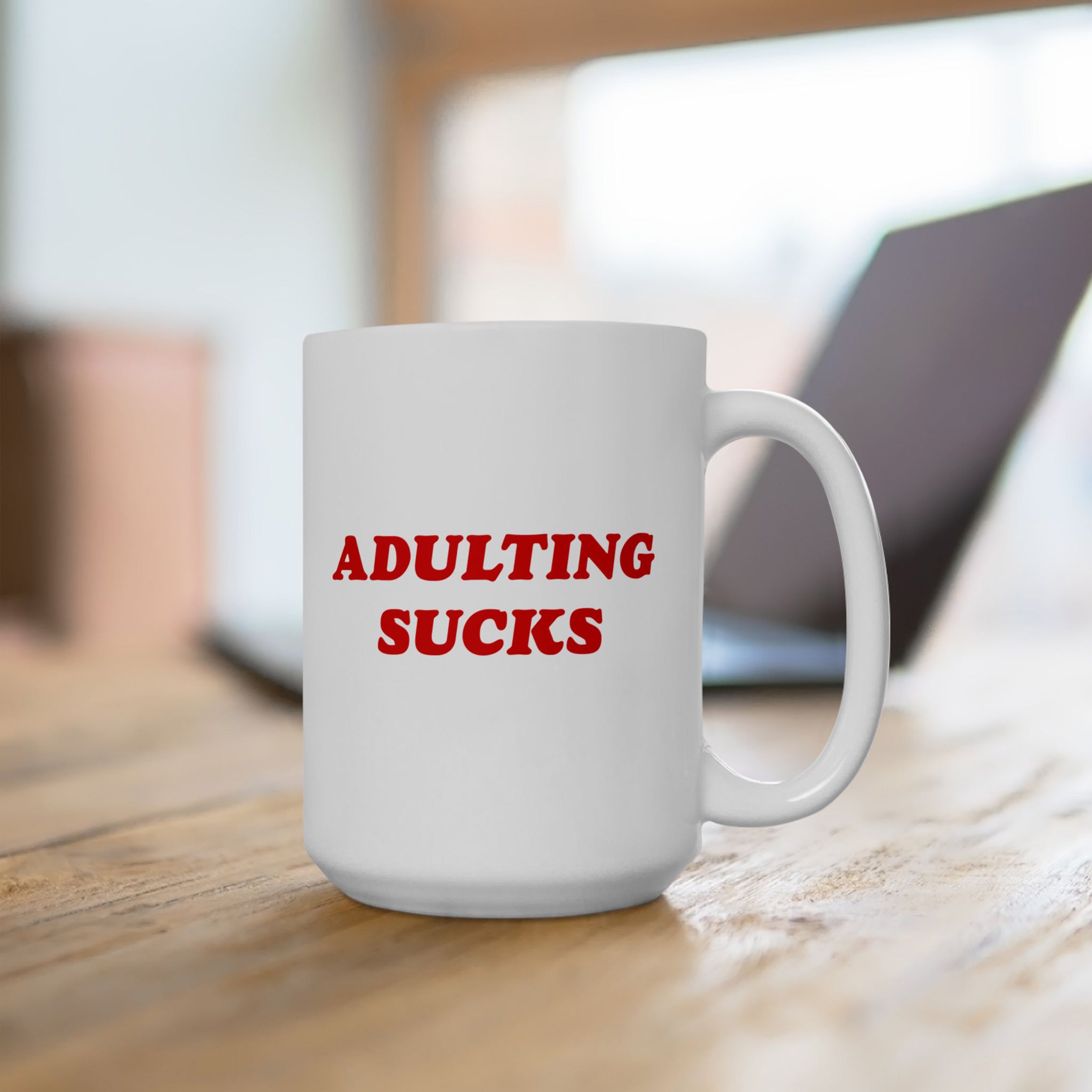 “Adulting Sucks” mug on a wood desk in front of a laptop, perfect for office sarcasm and remote work burnout