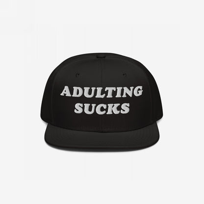 Black flat-brim snapback hat with ADULTING SUCKS embroidered in bold white letters, reflecting a candid, humorous take on life's challenges. Perfect for expressing shared frustrations while supporting a meaningful cause.