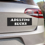 Close-up of “Adulting Sucks” sticker on a car bumper, showing bold white text on matte black background