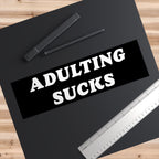 Flat lay of “Adulting Sucks” sticker with ruler for scale, surrounded by desk items to hint at workplace misery