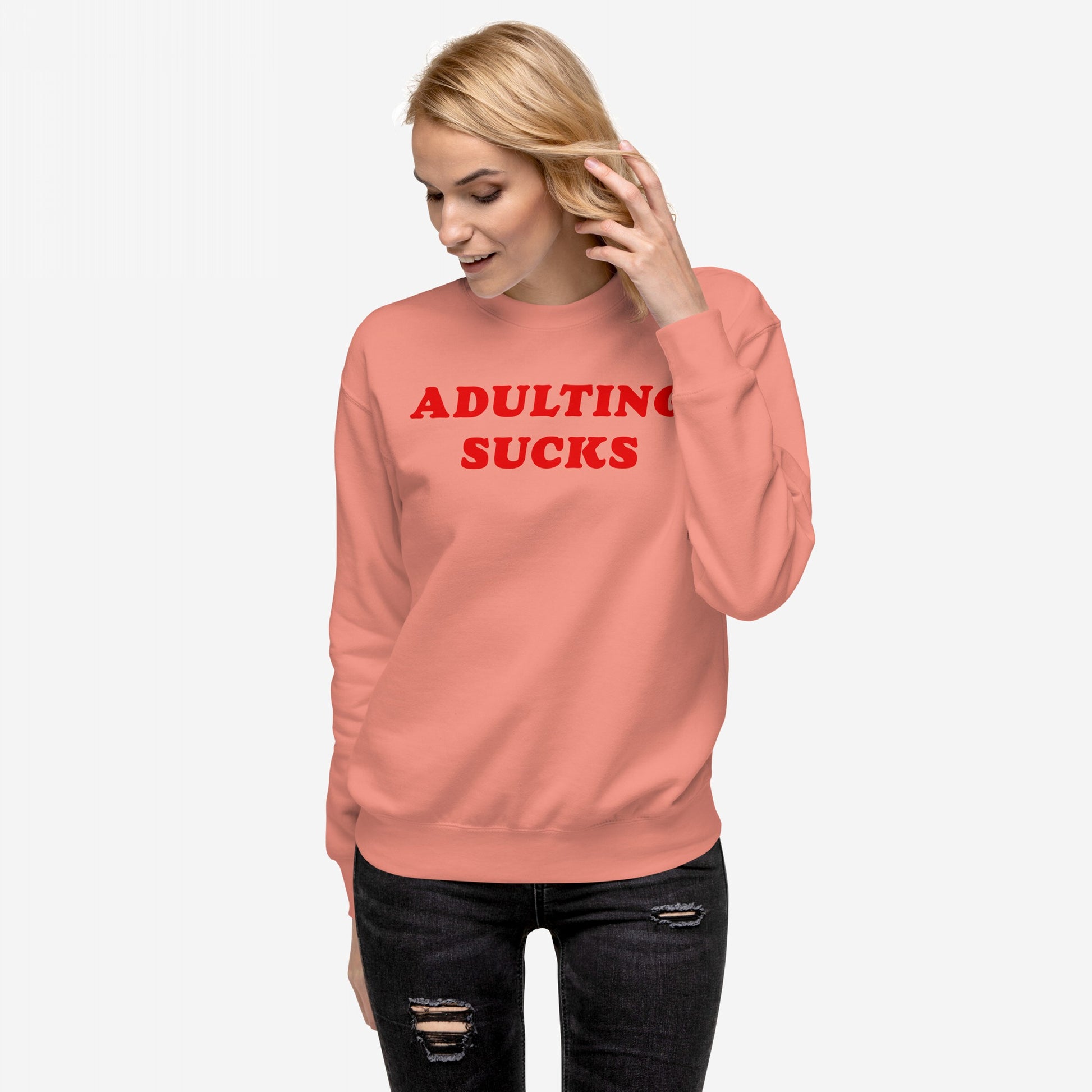 A young woman models the Adulting Sucks Hoodie & Sweatshirt, featuring bold red lettering on coral fleece, capturing a humorous take on the challenges of adulthood with stylish comfort.